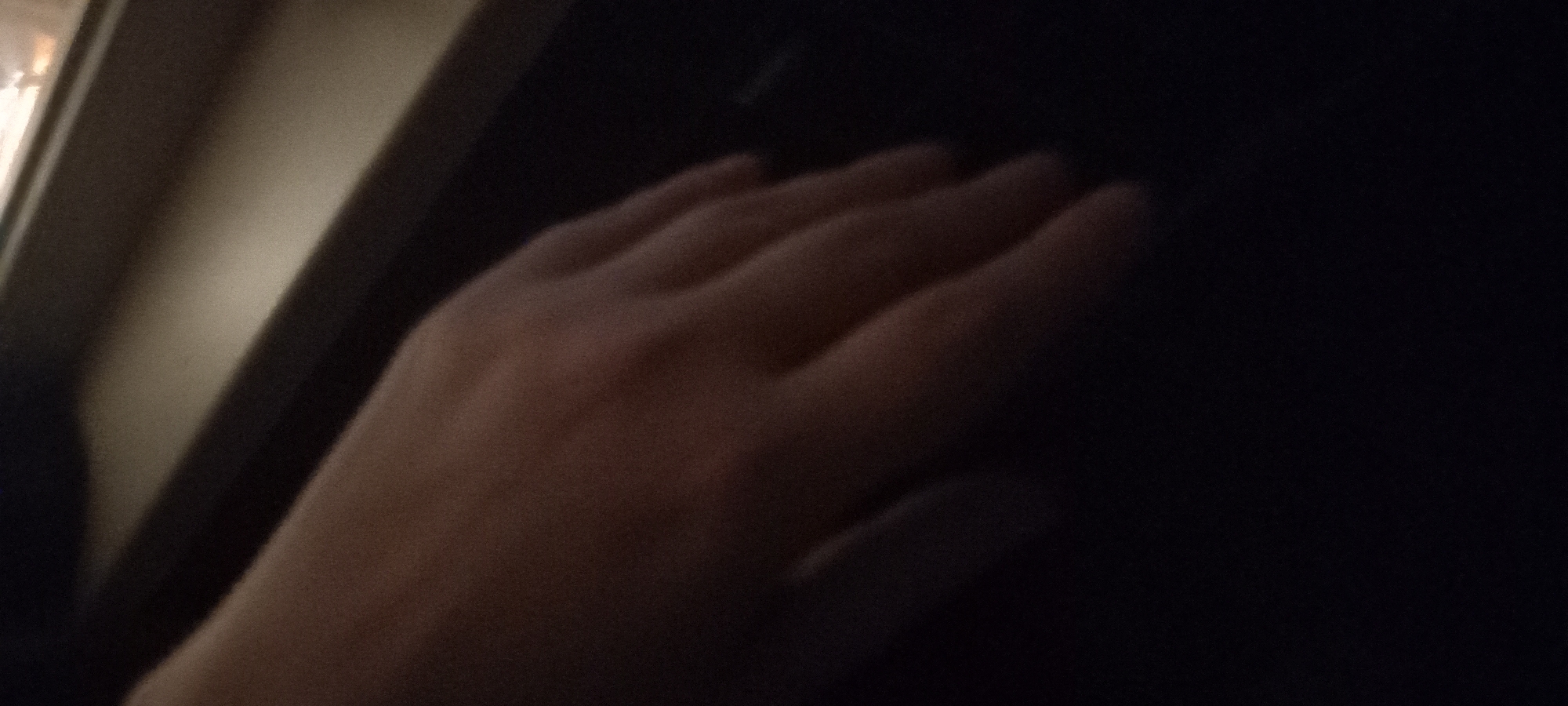 The image shows a close-up view of a person's hand, with the fingers gently curved. The hand is illuminated from the right, casting shadows that highlight the contours of the fingers and the back of the hand. The background is dark, with a faint suggestion of a lighter surface or object in the upper right corner. The scene appears to be indoors, given the lighting and the indistinct background. The setting does not provide any specific visual cues to pinpoint the location within Kryvyi Rih, Ukraine.