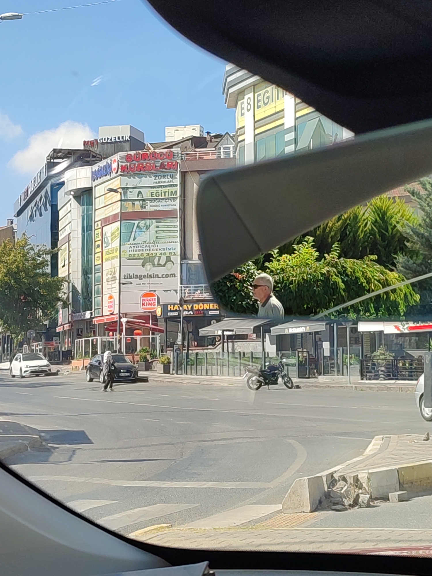 The image captures a street scene in Üsküdar, Turkey, likely during daytime. In the foreground, the reflection of a car's interior, possibly a dashboard or windshield, frames the view. The midground shows a street with a pedestrian crossing. Several vehicles are present: a white car is parked on the left, a dark-colored sedan is in motion on the street, and a motorcycle is parked on the right side of the road. A person is walking across the street on the pedestrian crossing.
The background features a multi-story building with various businesses advertised on its facade. Visible signs include "GÜZELLİK" (Beauty), "KURSLARI" (Courses), "EĞİTİM" (Education), and "Burger King". Another building further back displays "E8 EGİTİM". Green trees and foliage are visible behind the buildings, suggesting a relatively urban but green environment. The sky is blue with some wispy clouds. The overall impression is a typical urban street view with commercial activity.