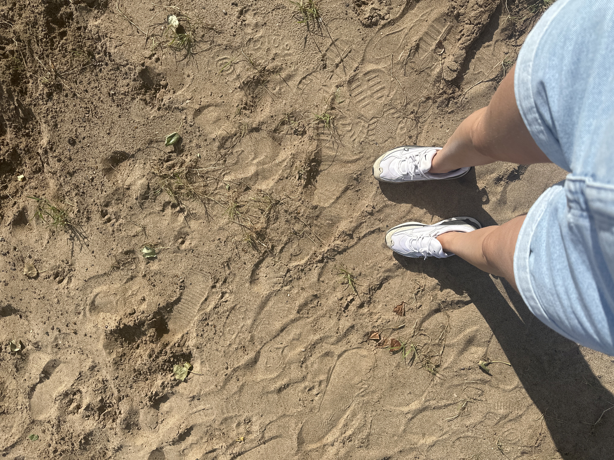 A person stands on sandy ground, their feet clad in white athletic shoes and light blue denim shorts. The sand is marked with numerous footprints, indicating previous foot traffic. Sparse green vegetation and dried leaves are scattered across the sandy surface. A long shadow stretches from the person, cast by an unseen light source, suggesting it is either morning or late afternoon. The context of Chaikovsky, Russia, implies this scene might be from a park, beach, or outdoor recreational area within the city, given its geography and typical amenities.