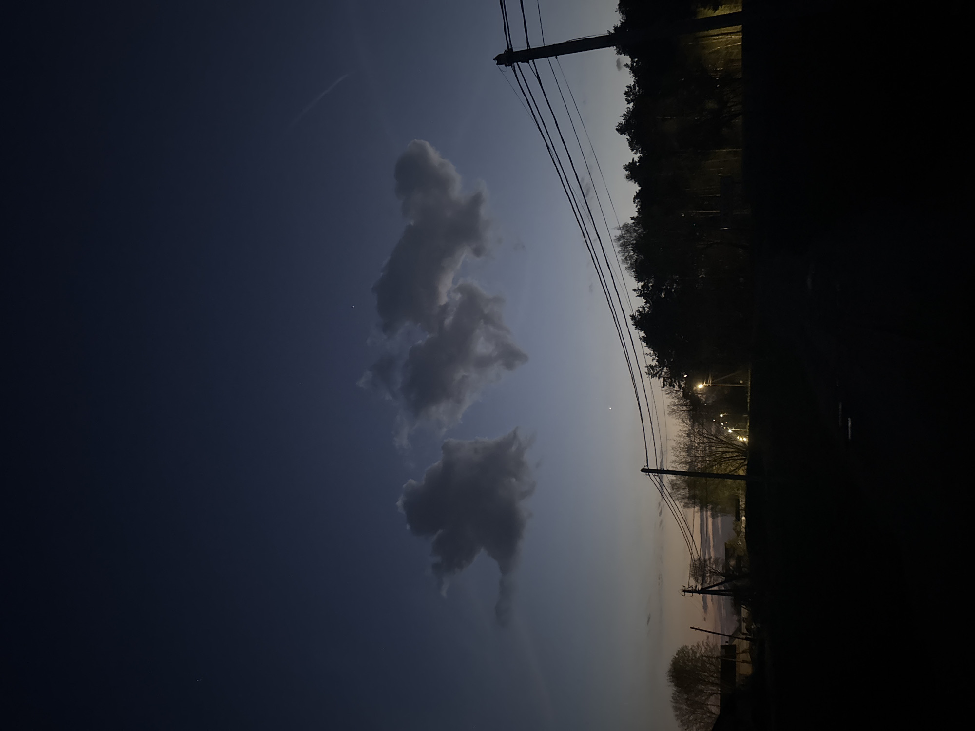 The image depicts a nighttime scene, likely in Раздолье, Russia, showing a section of the sky and a silhouetted foreground. The sky transitions from a dark blue on the left to a lighter, twilight hue on the right. Several puffy clouds are visible in the center of the frame. Power lines stretch across the upper right of the sky, connected to poles. The right side of the image shows a dark, undefined area likely representing trees and ground, illuminated by streetlights in the distance. The image orientation is unusual as it appears to be rotated 90 degrees counterclockwise.