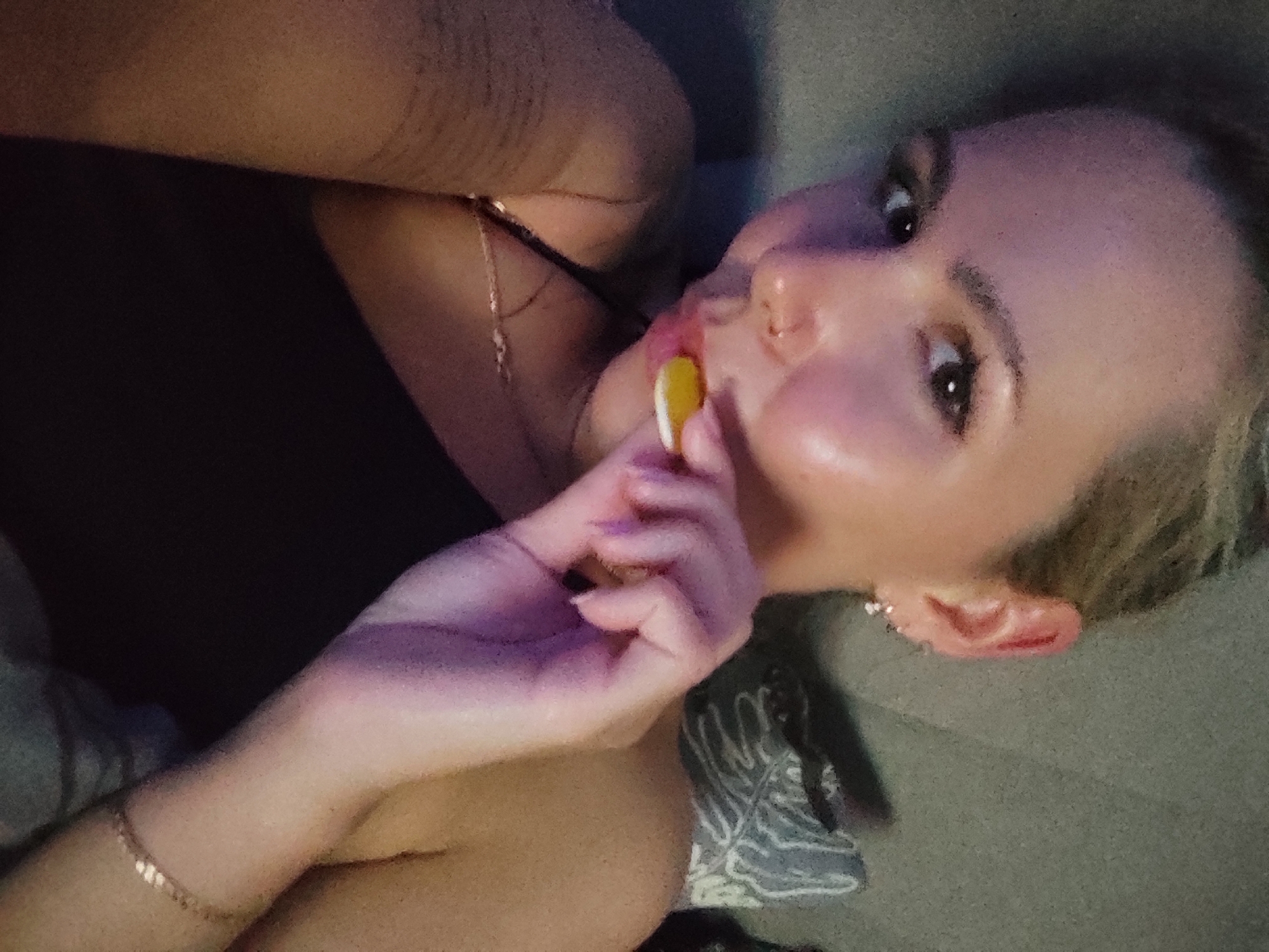 A woman lies on a gray surface, possibly a couch or bed. She is holding a yellow and white disc-shaped object near her mouth. She has light skin, light-colored hair, and is wearing a dark top. A gold chain necklace is visible around her neck. There is a patterned fabric or pillow near her shoulder. The image does not contain enough information to determine a location in Chaikovsky, Russia.