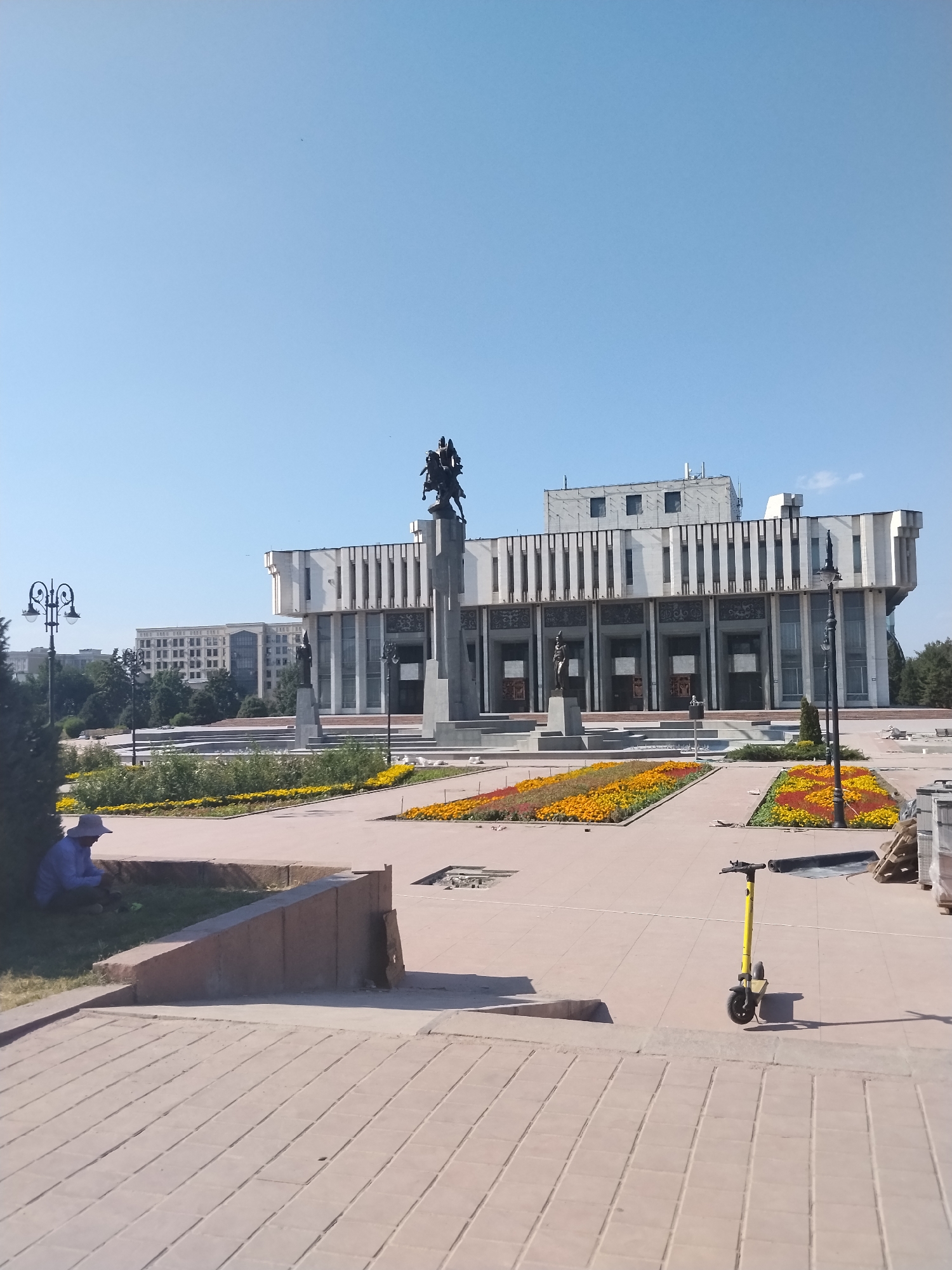 The image captures a wide shot of a public square in Bishkek, Kyrgyzstan, under a clear blue sky. Dominating the center background is a grand, modern building with a white facade, vertical window elements, and decorative panels. In front of this building stands a prominent monument featuring a large stone pedestal topped with a statue of two figures on horseback. To the left of the main building, another, slightly older-looking building with multiple windows is visible.
In the foreground, a person wearing a wide-brimmed hat and a light blue shirt is seated under a tree on the left side of the frame, appearing to be looking at their phone. The square itself is paved with light-colored tiles. Flowerbeds with vibrant yellow and orange flowers are arranged in rows, flanking the pathways. A yellow electric scooter is parked on the right side of the paved area. Streetlights with ornate designs are placed at intervals within the square. The scene suggests a public space that is well-maintained and likely a popular gathering spot.