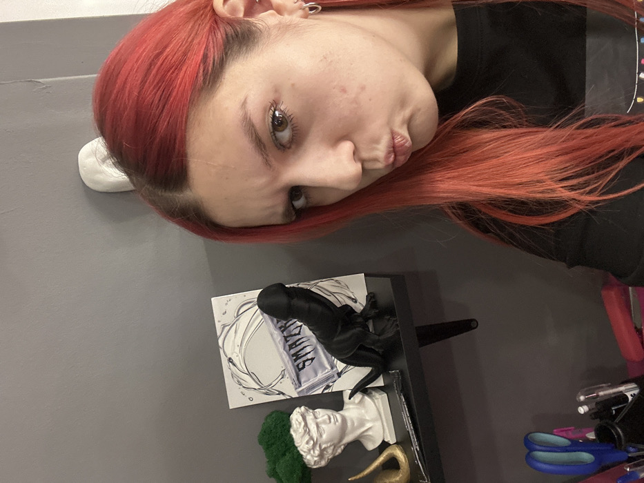 The image depicts a female individual with red-dyed hair in an indoor setting, identified as Saratov, Russia. She is positioned on the right side of the frame, from the forehead to the upper chest, looking directly at the camera with pursed lips and slightly furrowed eyebrows. She wears a black long-sleeved top and multiple silver hoop earrings in her visible left ear. A faint scar is present on her forehead, and minor blemishes are visible on her cheek.
The background consists of a solid dark grey wall. A black floating shelf is mounted on this wall. On the shelf, from left to right, are: a white bust resembling a classical male figure, topped with green moss-like material; a partially visible golden, curved object behind the bust; and a white square print with a water-splash design and the partially obscured stylized text "SMAZY". In front of the print stands a black figurine, composed of a dinosaur's body (resembling a Tyrannosaurus Rex) with a large, distinctively phallic head.
Below the shelf, out of clear focus in the lower-left, a pink container holds various writing instruments and a pair of blue-handled scissors. A red object is also partially visible in this area. A small white object, possibly a light switch or fixture, is affixed to the wall above the individual's head. The scene is evenly lit.