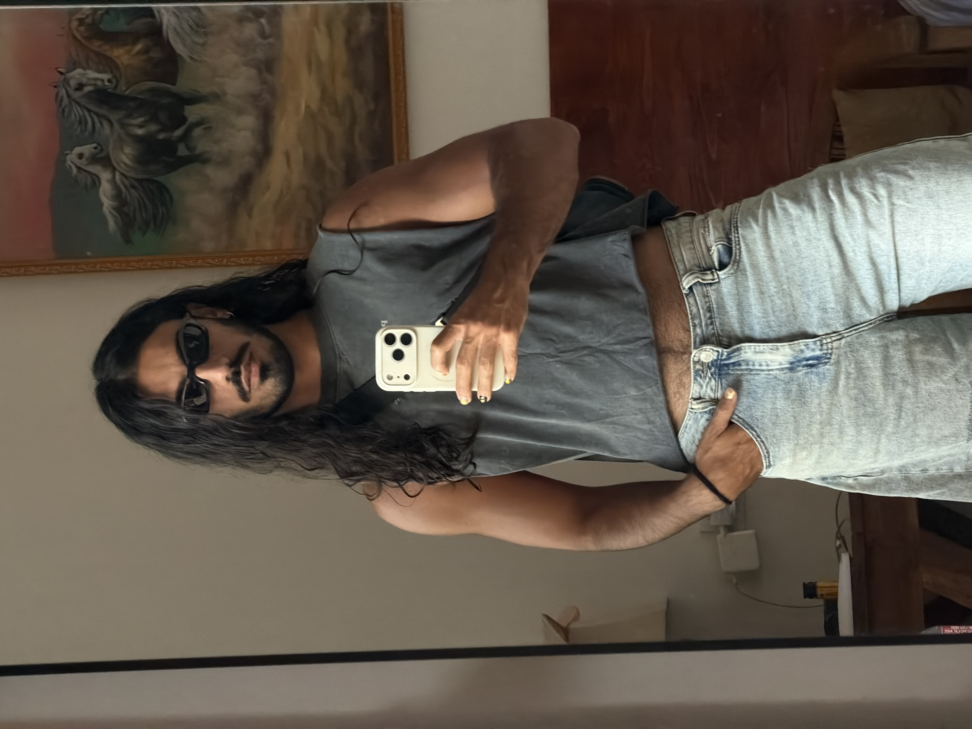 A person with long, dark hair and a goatee is taking a mirror selfie. They are wearing a dark gray, sleeveless, cropped t-shirt and light-wash, wide-leg jeans. They have on black sunglasses and are holding a white smartphone in their right hand, taking the photo. The background includes a large framed painting of horses on the right and a mirror on the left, reflecting the person and their surroundings. A wooden headboard and a glimpse of a wall are visible behind the person. The setting appears to be a bedroom.