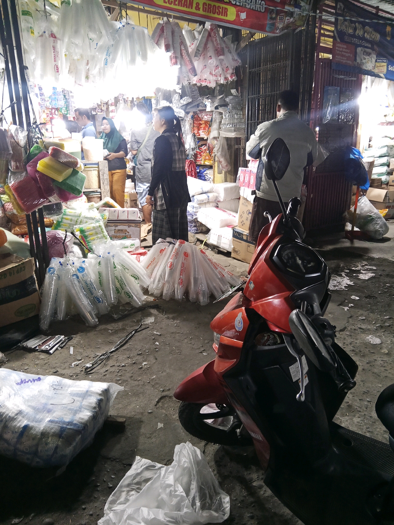The image depicts a street market scene in Lubuklinggau, Indonesia, likely a vendor stall. In the foreground, a red and black scooter is parked. Behind the scooter, a variety of goods are displayed. Hanging from the ceiling are numerous clear plastic bags, some containing what appear to be disposable cups or food containers. On a table or display shelf, stacks of colorful items, possibly plastic wares, are visible. Several people are present in the background, including a woman in a headscarf and another woman wearing a plaid shirt, seemingly interacting with the vendor or browsing the goods. The ground is dirty, with debris scattered around, suggesting an outdoor market environment. The overall impression is of a busy commercial area with a focus on disposable plastic products.