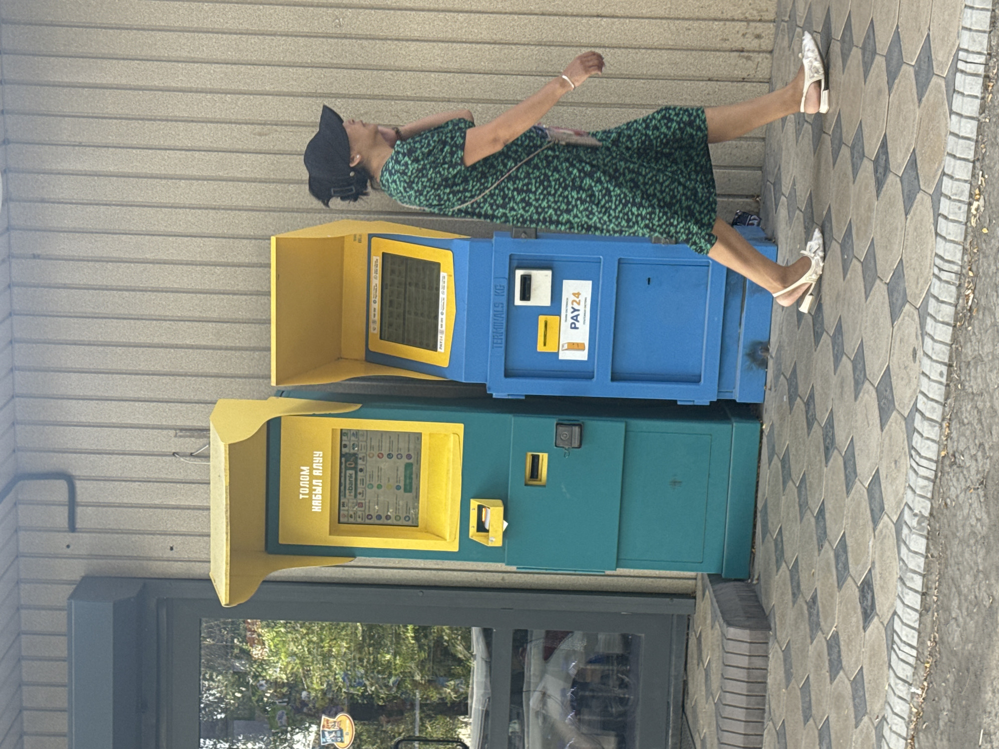A full-shot, eye-level, outdoor photograph captures a woman walking past two payment terminals in Bishkek City, Kyrgyzstan. The woman, wearing a green floral print dress and a black baseball cap, is captured in profile as she walks from right to left, speaking on her mobile phone. Her white, floral-patterned flat shoes and a small crossbody bag complete her attire.
To her left, two payment terminals stand adjacent to each other. The terminal on the left is predominantly teal with a yellow hood and a yellow touchscreen display. The screen shows various icons and text, including "ТОЛОМ НАВЫЯ АЛУЧ" and "Hbank". Below the screen, there is a slot for cash or cards, and a smaller yellow receptacle.
The terminal on the right is primarily blue with a yellow hood and a blue touchscreen display. The screen shows text and icons, including "PAY24". Below the screen are a card reader slot and a white rectangular panel. Text on the blue casing reads "TERMINALS KG".
The terminals are situated on a patterned tiled sidewalk with a border of smaller bricks. The background consists of a light-colored, ribbed wall. To the far left of the image, a portion of a glass door and the reflection of trees and a white car are visible. The lighting suggests it is daytime, with sunlight casting some shadows.