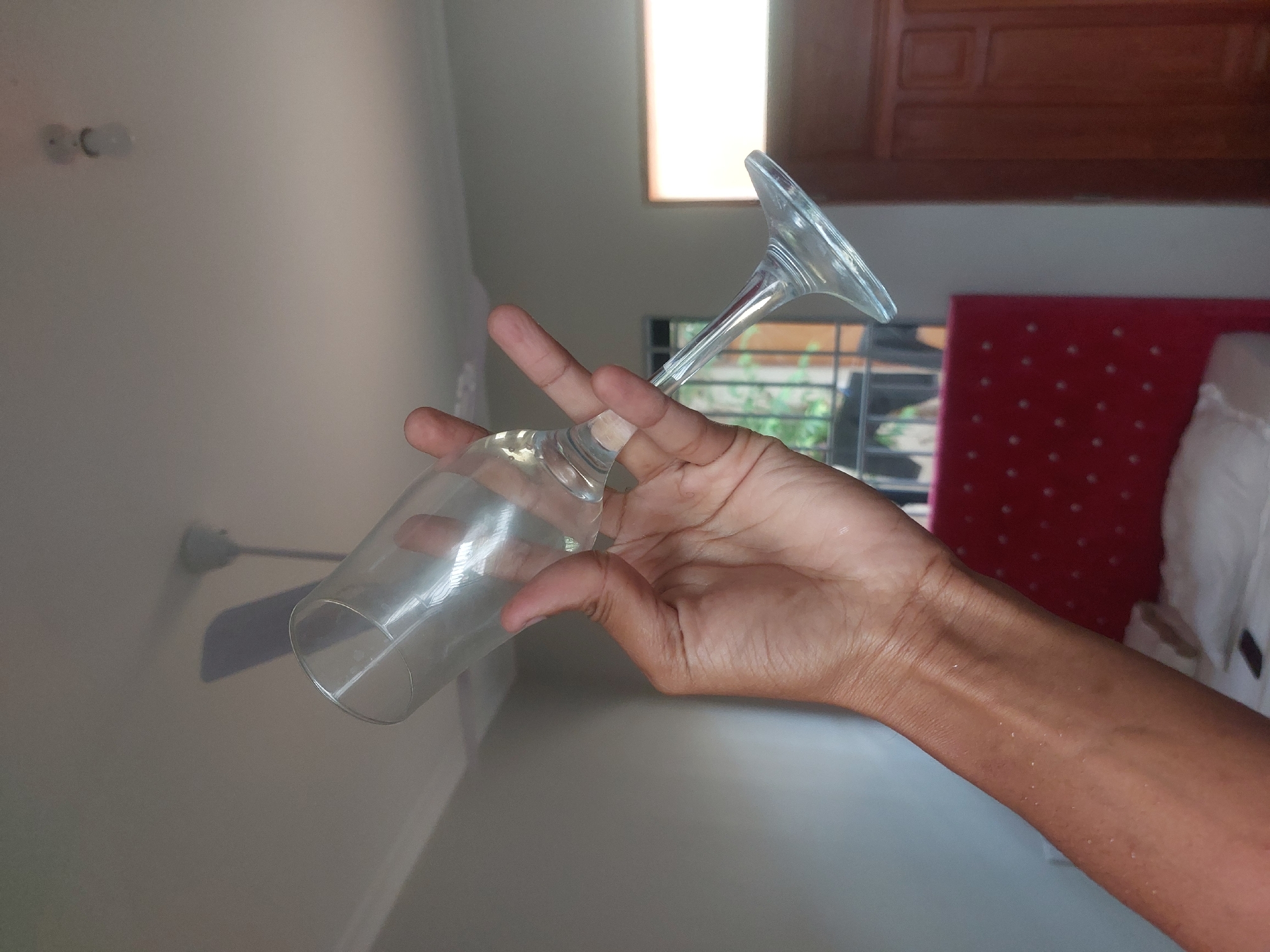 A right hand is holding a clear glass wine glass at an angle. The glass is empty and tilted downward. The background includes a wall, a window with a view of green vegetation, a red headboard, and a white pillow. A ceiling fan is also visible in the background. The location appears to be indoors, possibly a residential room. It is not possible to ascertain the location as Dar es-Salaam, Tanzania from the available imagery.