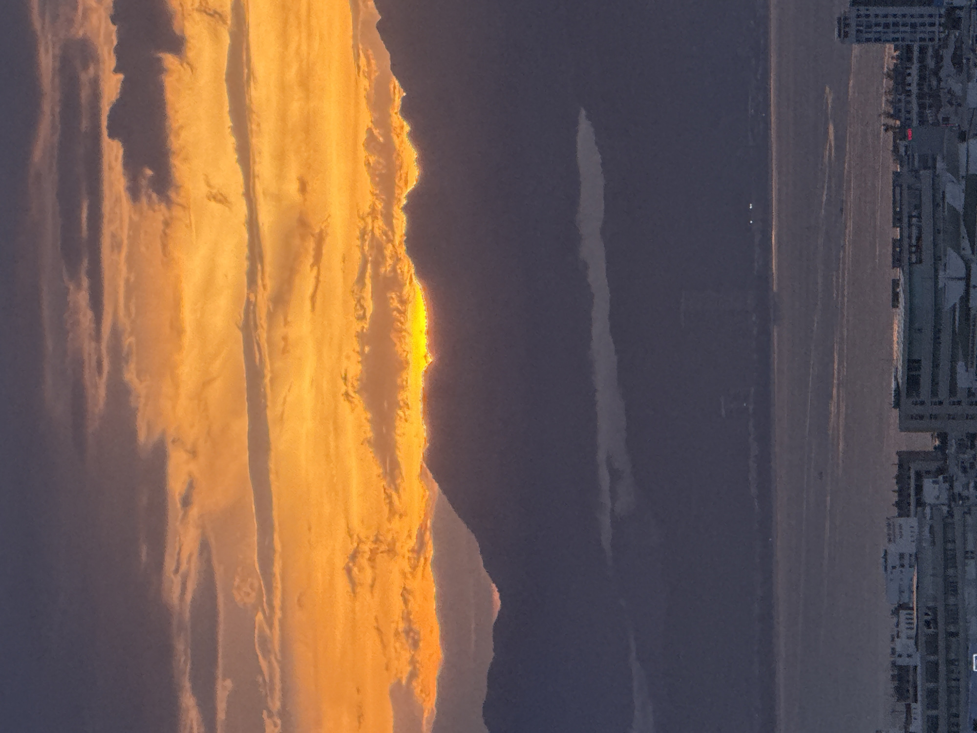 This image captures a sunset over the city of Da Nang, Vietnam. The sky is a dramatic display of orange and purple clouds, with the sun just beginning to dip below the horizon, casting a warm glow. In the foreground, a cityscape of buildings is visible, and beyond that, mountains form a silhouette against the vibrant sky. The ocean stretches out between the city and the mountains, reflecting the colors of the sunset. The scene is peaceful and serene, with the natural beauty of the sunset contrasting with the urban landscape.