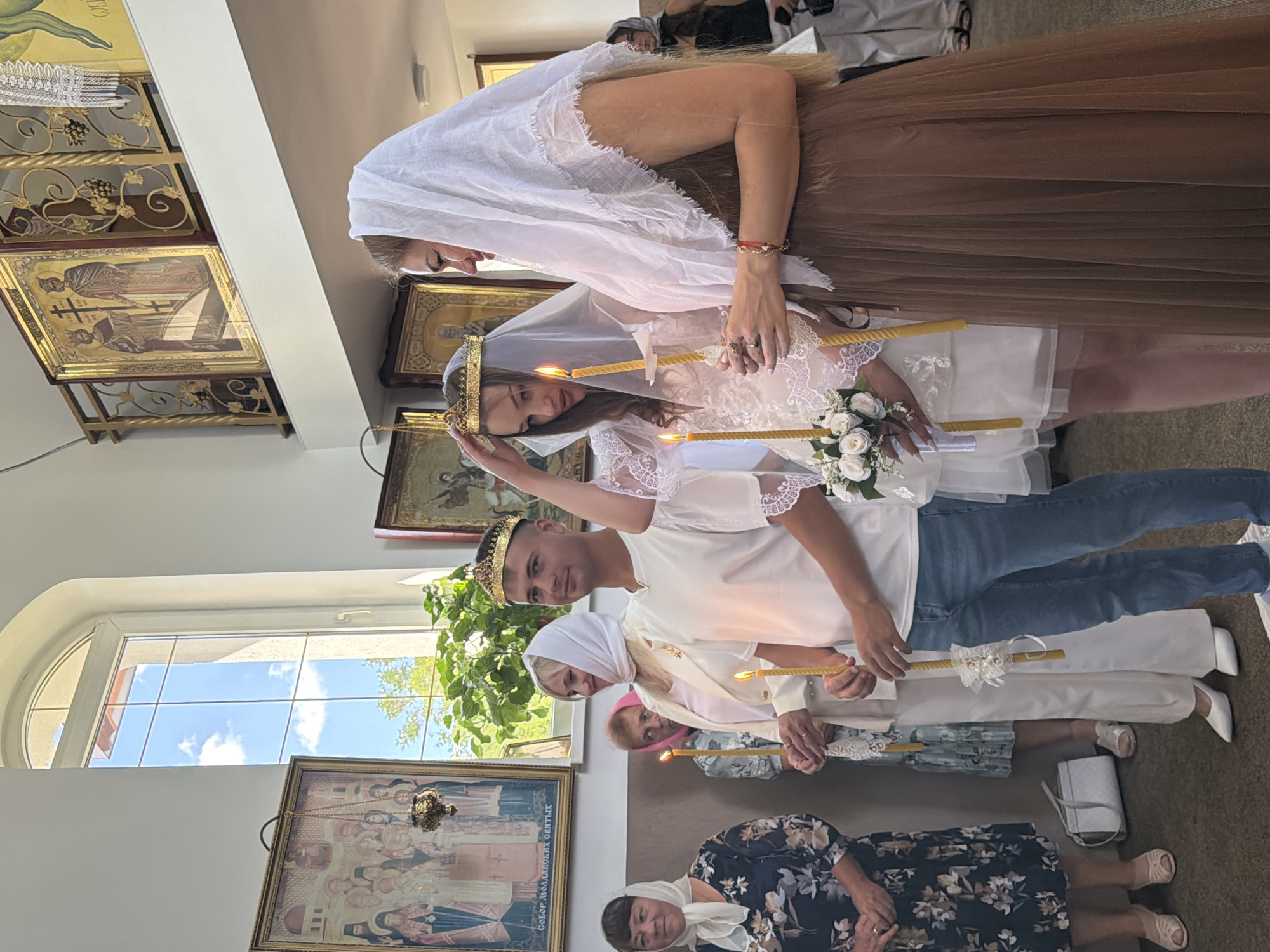 The image depicts an indoor religious ceremony, likely a wedding, taking place within a church in Strășeni, Moldova. The central figures are a young couple, appearing to be the bride and groom, both wearing crowns, a symbolic element of Orthodox wedding traditions. The groom is on the right, facing the camera, while the bride is to his left, looking down. They are both holding lit candles, with a small bouquet of white flowers positioned in front of the bride.
Flanking the couple are several other individuals. To the groom's left is a woman in a white suit and veil, also holding a candle. Further to the left, a woman in a patterned dress and headscarf stands with her hands clasped. Behind the bride, another woman in a pink headscarf is visible, also holding a candle. To the far right, partially obscured, is a woman in a brown dress and white headscarf, her back mostly to the camera. Further back, other attendees are present, including a woman in a black top and headscarf.
The environment appears to be a richly decorated church interior. Religious icons are visible on the walls, with one prominent icon featuring figures with halos. A large arched window with panes allows natural light to enter the space, illuminating the scene and revealing greenery outside. The flooring is a muted carpet. The overall atmosphere suggests a formal and solemn occasion.