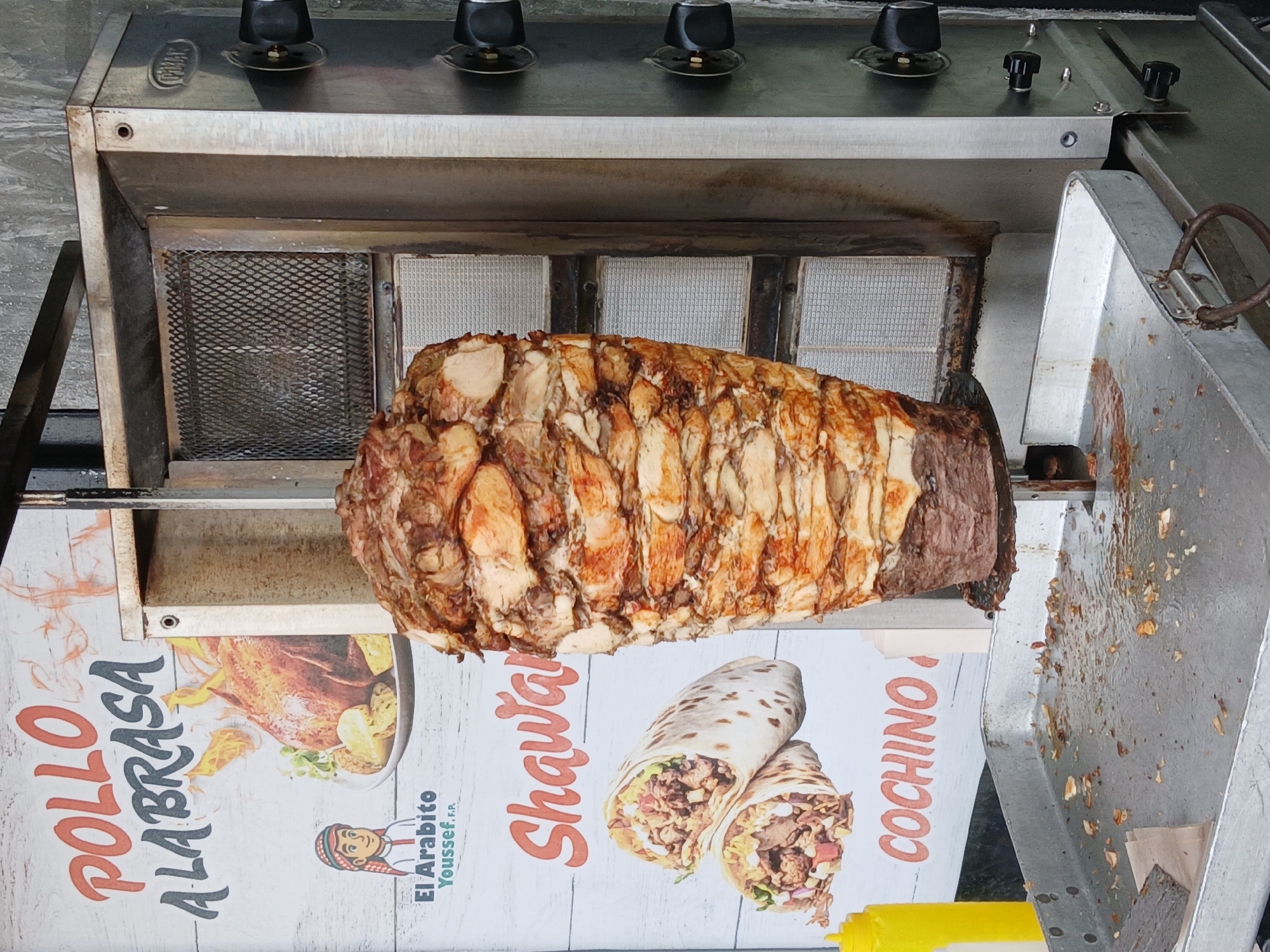 The image shows a vertical rotisserie for cooking meat, likely shawarma or similar. The rotisserie is made of stainless steel and has multiple burners at the top. A large cone of stacked meat is mounted on a vertical spit, positioned in front of the heating elements. The front of the rotisserie has promotional signage featuring images of roasted chicken ("Pollo A La Brasa"), shawarma wraps, and the word "COCHINO" (Spanish for "pork/pig"), alongside the name "El Arabito Youssef F.P.". Based on the provided information, this setup is located in Ciudad Guayana, Venezuela.