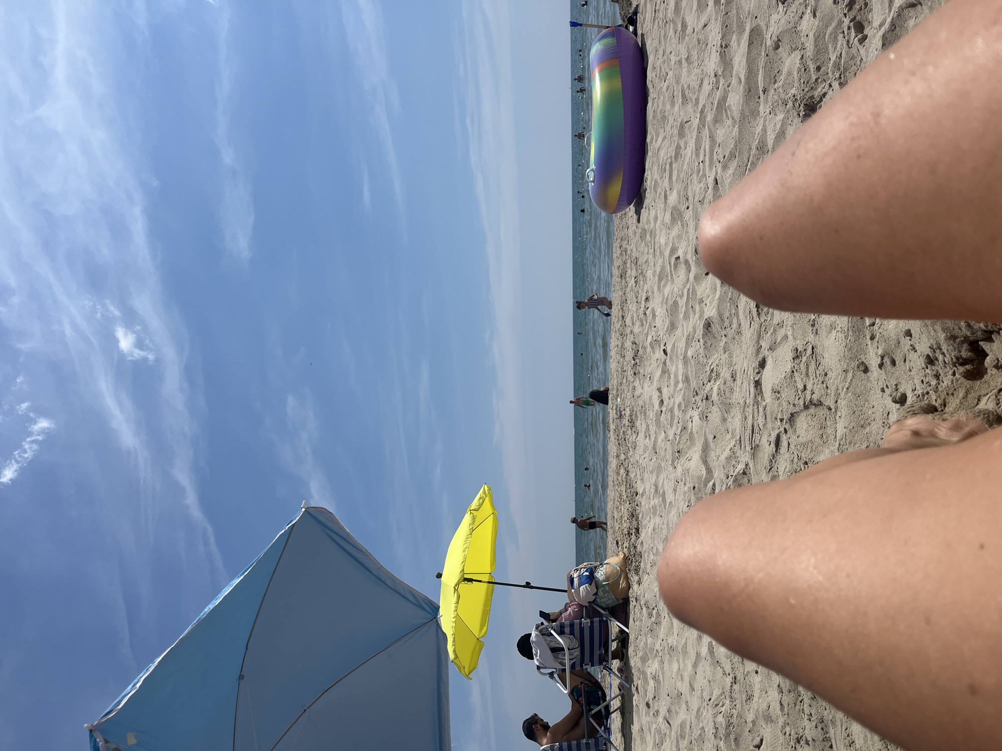 The image depicts a sunny day at a beach, likely Palavas-les-Flots, France, based on the provided context. In the foreground, a person's legs are visible, resting on the sandy beach.
Behind the legs, several beach umbrellas provide shade. A large blue umbrella is partially visible on the left, and a bright yellow umbrella is open, under which two people are seated in chairs. One person is looking at their phone, while the other is facing away. Beach bags and towels are also present near the chairs.
The sandy beach stretches towards the sea, where several individuals are enjoying the water. Some people are swimming or wading, while a child is visible on the shore with a toy. In the mid-ground, a large, colorful inflatable ring lies on the sand, with a shovel stuck in the sand beside it.
The sea is calm, with a gentle horizon line where it meets the sky. The sky is a clear blue with scattered white clouds, indicating fair weather. The overall scene conveys a relaxed and leisurely atmosphere of a typical summer day at the beach.