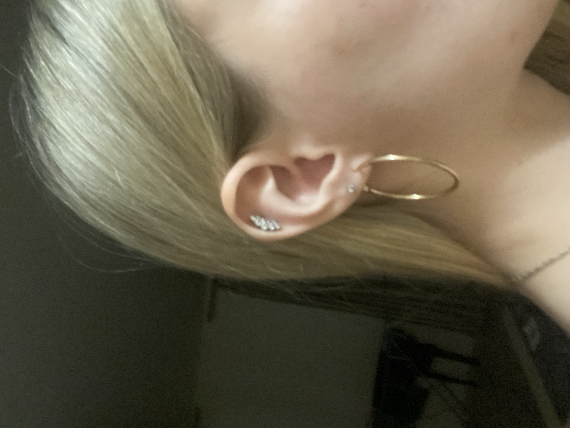 A close-up, slightly angled shot focuses on a person's ear and surrounding areas. The ear is adorned with a gold hoop earring and several small, possibly diamond or rhinestone-encrusted studs along the outer rim. The skin appears fair, and the person has blonde hair. Part of a necklace chain is also visible in the frame. The image background is blurry and dark. Location information is unavailable.