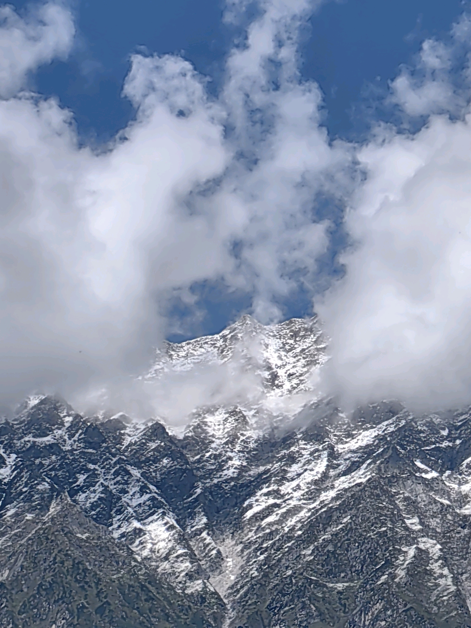 The image depicts a close-up view of snow-capped mountains. The sky is a vibrant blue with scattered white clouds. The mountains themselves are rugged and appear to be made of rock with patches of snow covering their peaks and crevices. The texture of the rock is rough, with various shades of gray and brown visible. The snow is white and contrasts sharply with the darker rock formations. The clouds are dynamic, with some obscuring parts of the mountain peaks, creating a sense of depth and atmosphere. The overall impression is one of a dramatic and majestic natural landscape.