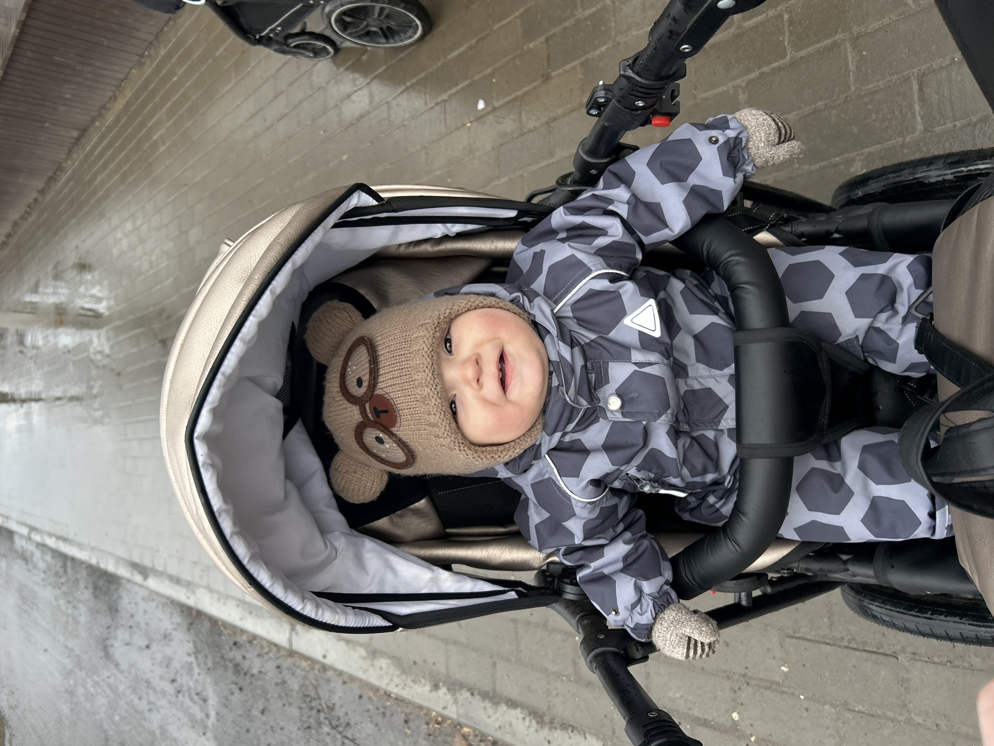 A baby wearing a brown bear hat and a gray patterned snowsuit is sitting in a stroller. The stroller has a light tan exterior and a white interior. The stroller is on a gray paved surface, possibly a sidewalk, which appears wet. Another stroller is partially visible in the upper left corner of the image. The image was captured on a wet day in what is likely Chaikovsky, Russia.