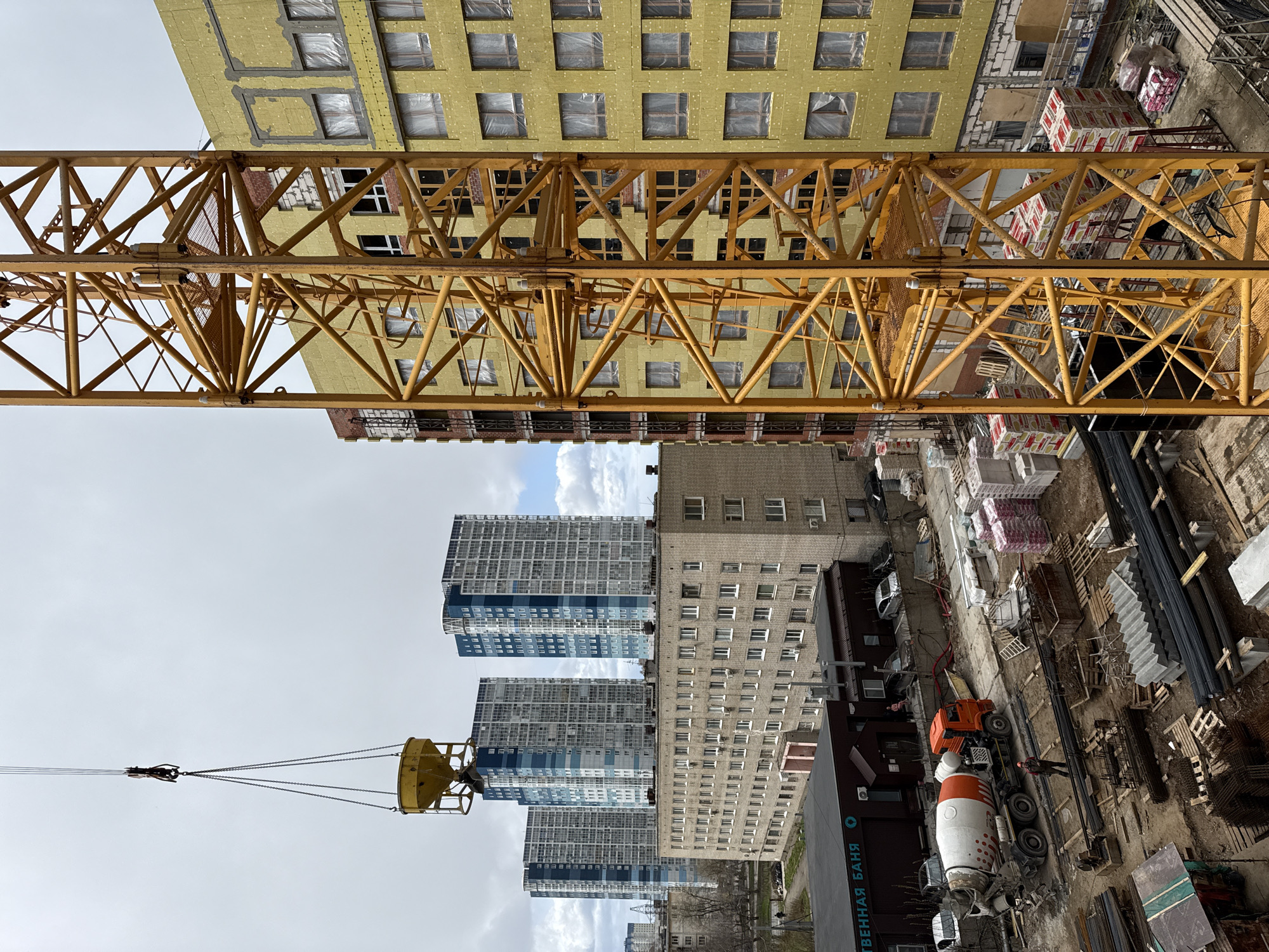 This image captures a vibrant construction site from an elevated perspective, likely from a crane or a nearby tall structure. The scene is set in an urban environment with a mix of old and new architecture.
Dominating the foreground is the robust yellow metal framework of a large tower crane's horizontal jib, spanning across the upper part of the frame. On the far left, a yellow concrete bucket is suspended from the crane's hook block by chains, suggesting material transport is underway or imminent.
In the mid-ground, a building under construction is prominent, clad in yellow insulation material, with numerous window openings covered in plastic sheeting. Beyond this, several modern, tall apartment buildings with blue and white facades stand out against an overcast sky. Older, brick-colored buildings with multiple windows are also visible.
On the ground level of the construction site, various materials are stacked, including pallets of bricks or blocks (some in red and white packaging), rebar, and wooden pallets. An orange and white concrete mixer truck is parked on the site, and a single person, possibly a construction worker in an orange vest, is visible near it.
The time of day appears to be daytime, with bright, diffused light from the cloudy sky. A notable detail providing a strong location cue is the text "ДЕРЕВЕННАЯ БАНЯ" (Derevennaya Banya), meaning "Village Bathhouse," visible on a dark-colored building at ground level, indicating a Russian-speaking country. The architecture of the tall residential buildings further supports this.