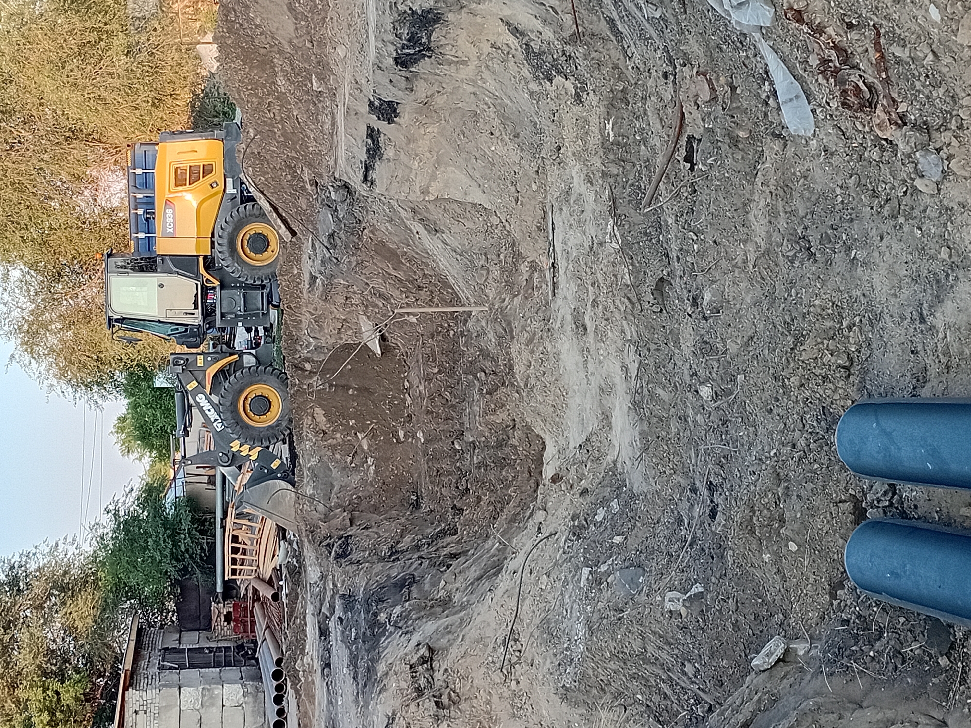 The image depicts a construction site with a yellow wheel loader labeled "XCMG XC936" parked on a pile of dirt. In the foreground, a deep excavation is visible, with various construction materials such as pipes and wooden planks scattered around. To the left, a brick building with a metal door stands adjacent to the site. The environment appears to be outdoors, with trees visible in the background under a clear sky. The scene is consistent with ongoing construction or excavation work in an urban or semi-urban setting, potentially in the city of Semey, Kazakhstan, given the provided context. There are no people visible in the image.