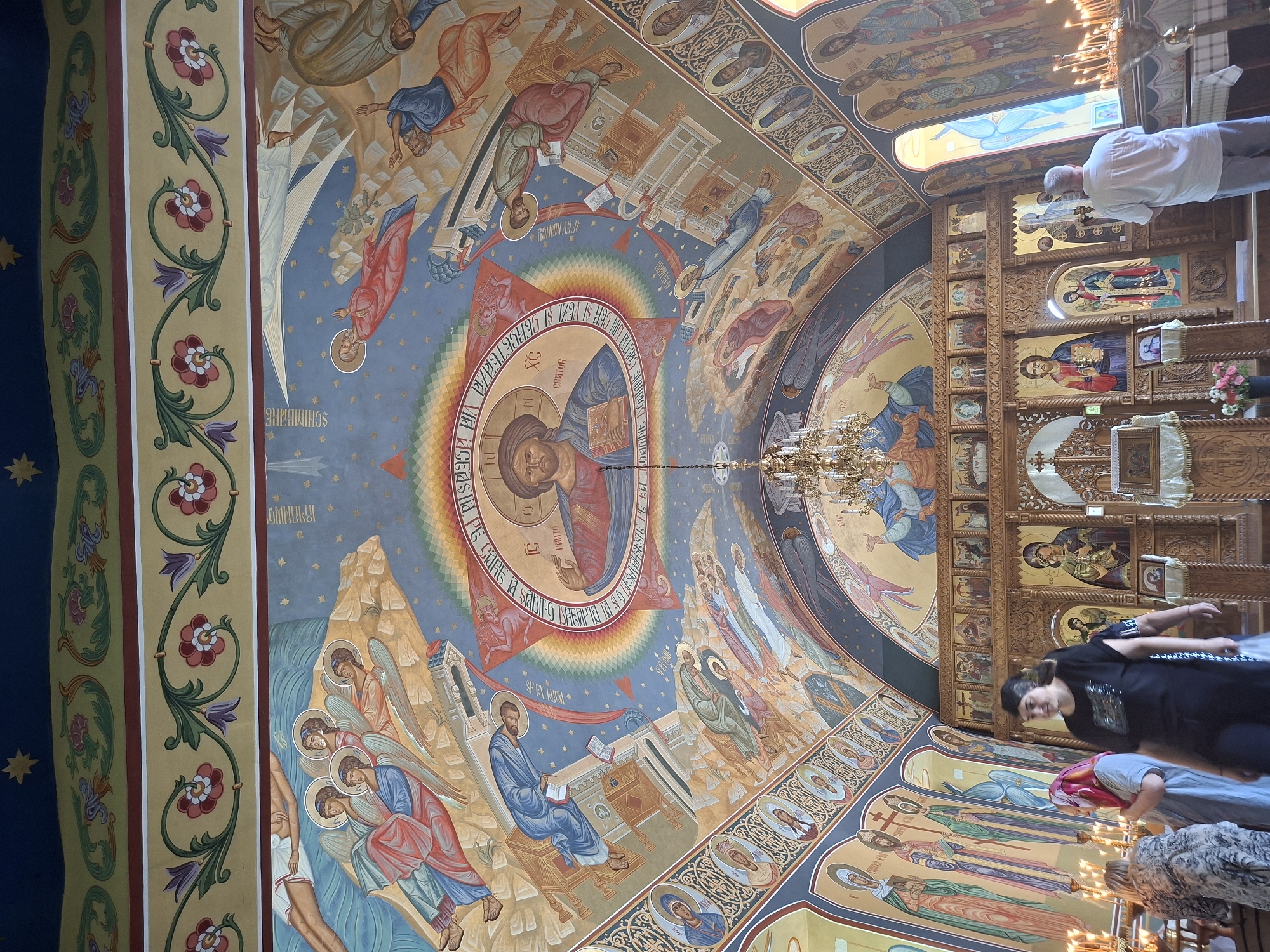 The interior of a church in Condrița, Moldova, features ornately painted ceilings and walls. The ceiling depicts religious scenes with figures like Jesus, angels, and other biblical characters rendered in vibrant colors. A large fresco of Jesus is centered in a circular motif on the ceiling, surrounded by a rainbow-colored halo and inscriptions. Below the ceiling, along the walls, are rows of painted icons of saints. The main focus in the foreground is the altar area, which includes an iconostasis, a screen or wall of icons, with several large, framed religious paintings. A chandelier hangs from the ceiling above the altar. Several individuals are present in the church. In the foreground, three women are walking or standing. Further back, a man is standing facing the altar, seemingly in prayer. Candles are lit on candelabra throughout the altar area, and floral arrangements are visible. The architecture includes arched windows and a coffered ceiling border with decorative floral patterns.