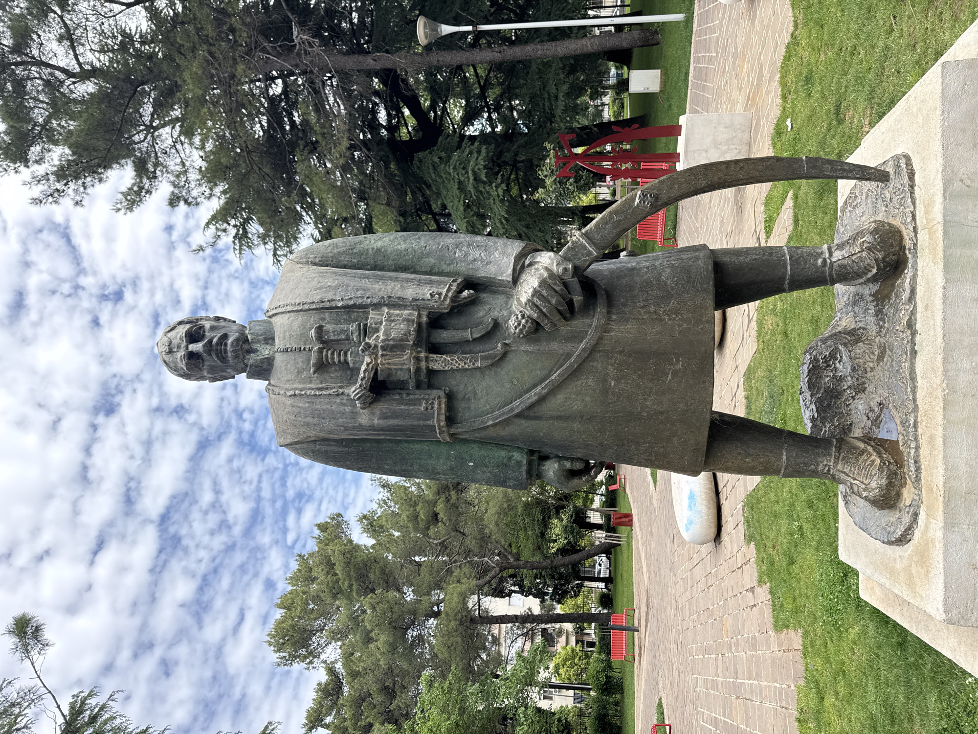 A bronze statue stands in a park in Podgorica, Montenegro. The statue depicts a man in historical clothing with a stern expression. He is holding an object resembling a scythe or a curved blade in his left hand. He is standing on a stone base on top of a concrete pedestal. Behind the statue are trees, a pathway made of brick, benches, and a red abstract metal sculpture. The sky above is cloudy.