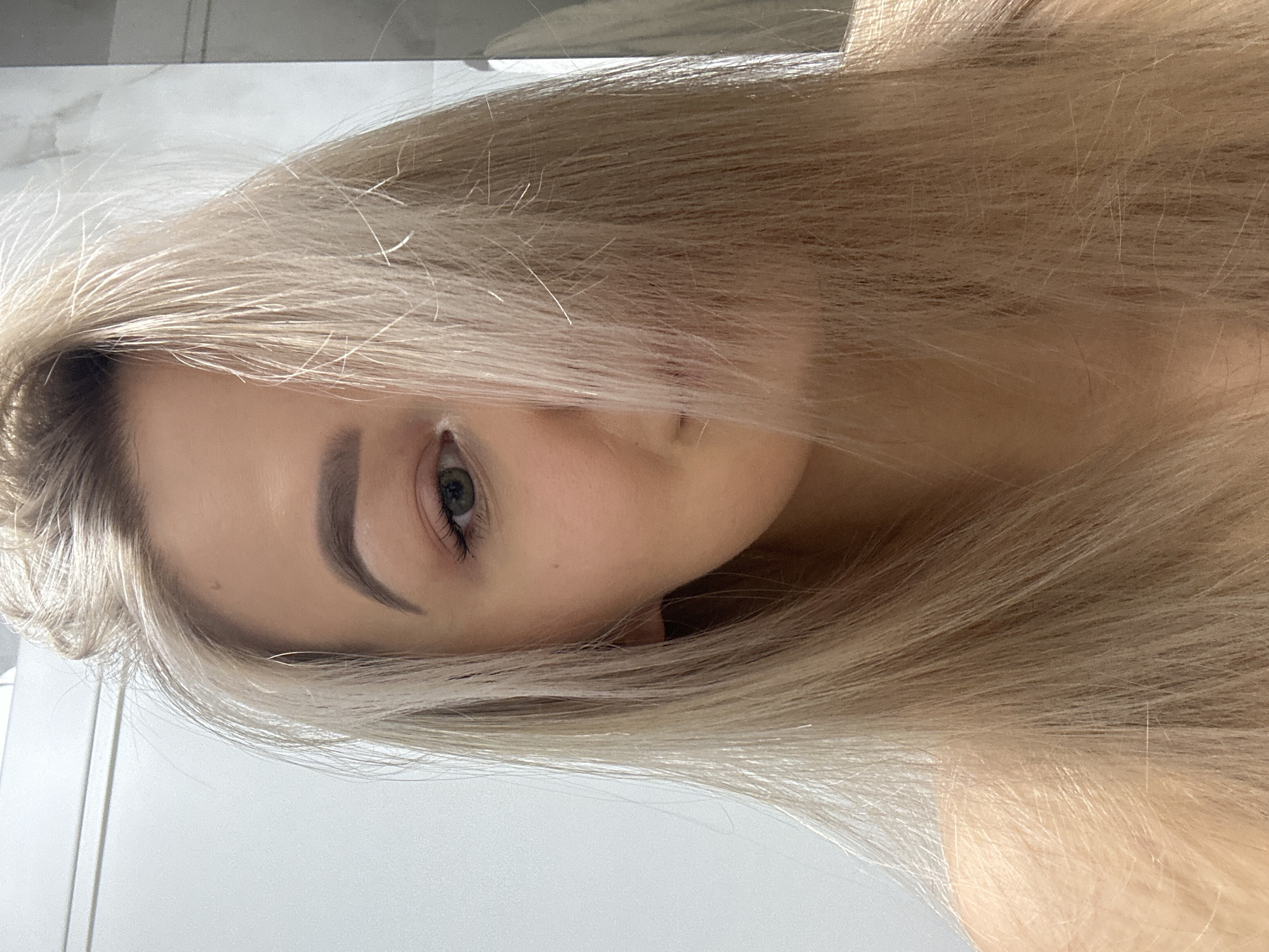 A close-up portrait shows a person with long, blonde hair partially obscuring their face. One eye is visible, and the background appears to be a white wall or surface. The image is rotated to the side. Location is not determinable from the visual information.