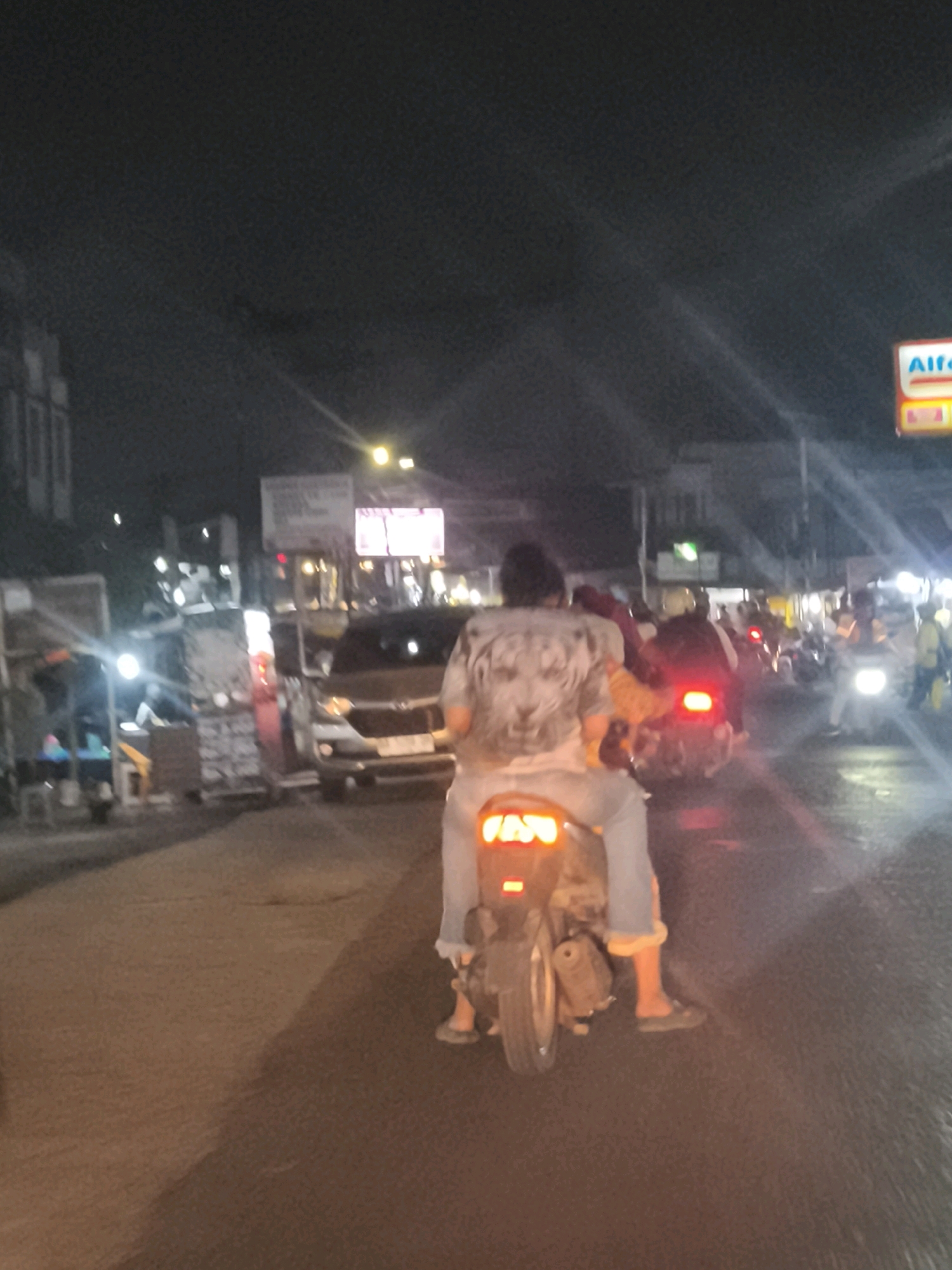 The image depicts a nighttime street scene in Palembang, Indonesia, likely on a humid evening given the atmospheric conditions. In the foreground, a person is riding a motorcycle with a passenger behind them. The rider is wearing a light-colored t-shirt with a graphic print, possibly a tiger's face. The passenger is wearing a dark top and appears to be holding onto the rider. The motorcycle has a prominent red taillight illuminated. To the left, a car is visible, and further back, there are streetlights and buildings, suggesting an urban environment. On the right side of the image, a red and white sign with the "ALFA" logo is visible, indicating a commercial establishment, possibly an Alfamart convenience store, which are common throughout Palembang. The road surface appears wet, reflecting the lights, which could be due to rain or recent cleaning. The overall impression is a typical busy street scene in Palembang at night, with traffic and commercial activity.