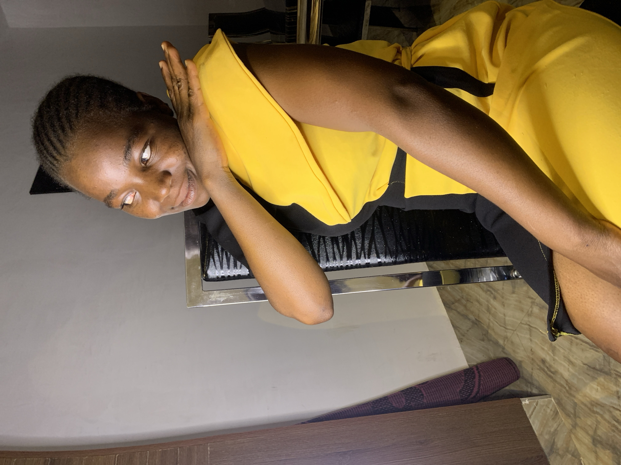 A woman is reclining on a black and silver chair in what appears to be an indoor space. She is wearing a yellow and black outfit. Her left hand is raised near her face, and she is looking slightly off-camera with a slight smile. The background includes a gray wall, a portion of a wooden structure (possibly a bed frame), and a rolled-up maroon and black mat on the floor. There is insufficient information to determine the location within the city of Jos, Nigeria.