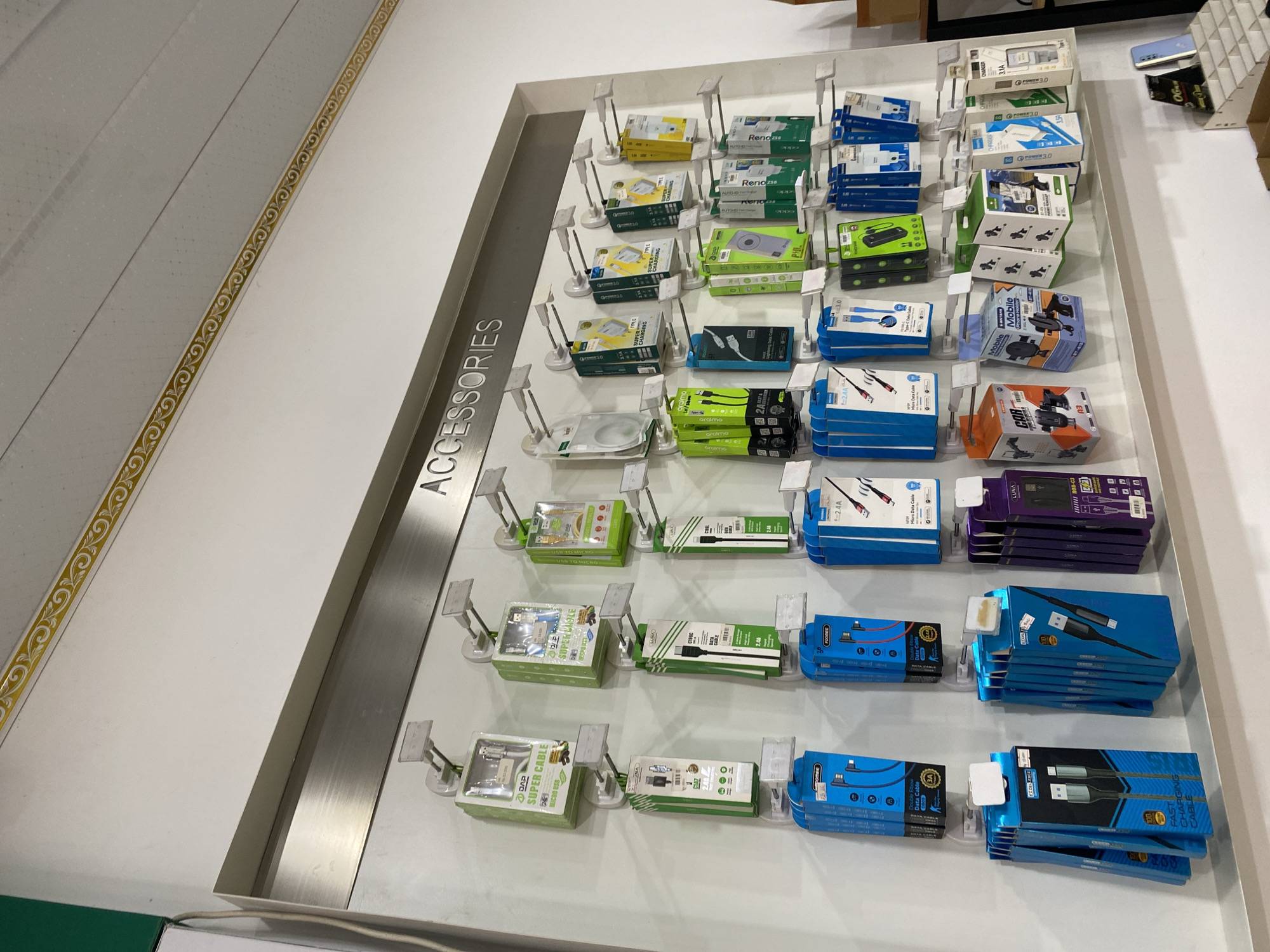 The image shows a retail store display of mobile phone accessories, specifically chargers and cables, in Ta, Indonesia. The accessories are organized on shelves and hooks against a white wall with a decorative border at the top. Several rows of products are visible, with many items packaged in colorful boxes. Brands like DAD, LUNA, oraimo, and Foomer are discernible on some packaging. The products are predominantly cables of various types (USB-C, Micro USB) and some phone holders. In the lower right corner, there is a white display stand holding a few items, and a box. The lighting appears to be artificial, typical of an indoor retail environment. There are no people visible in the image.