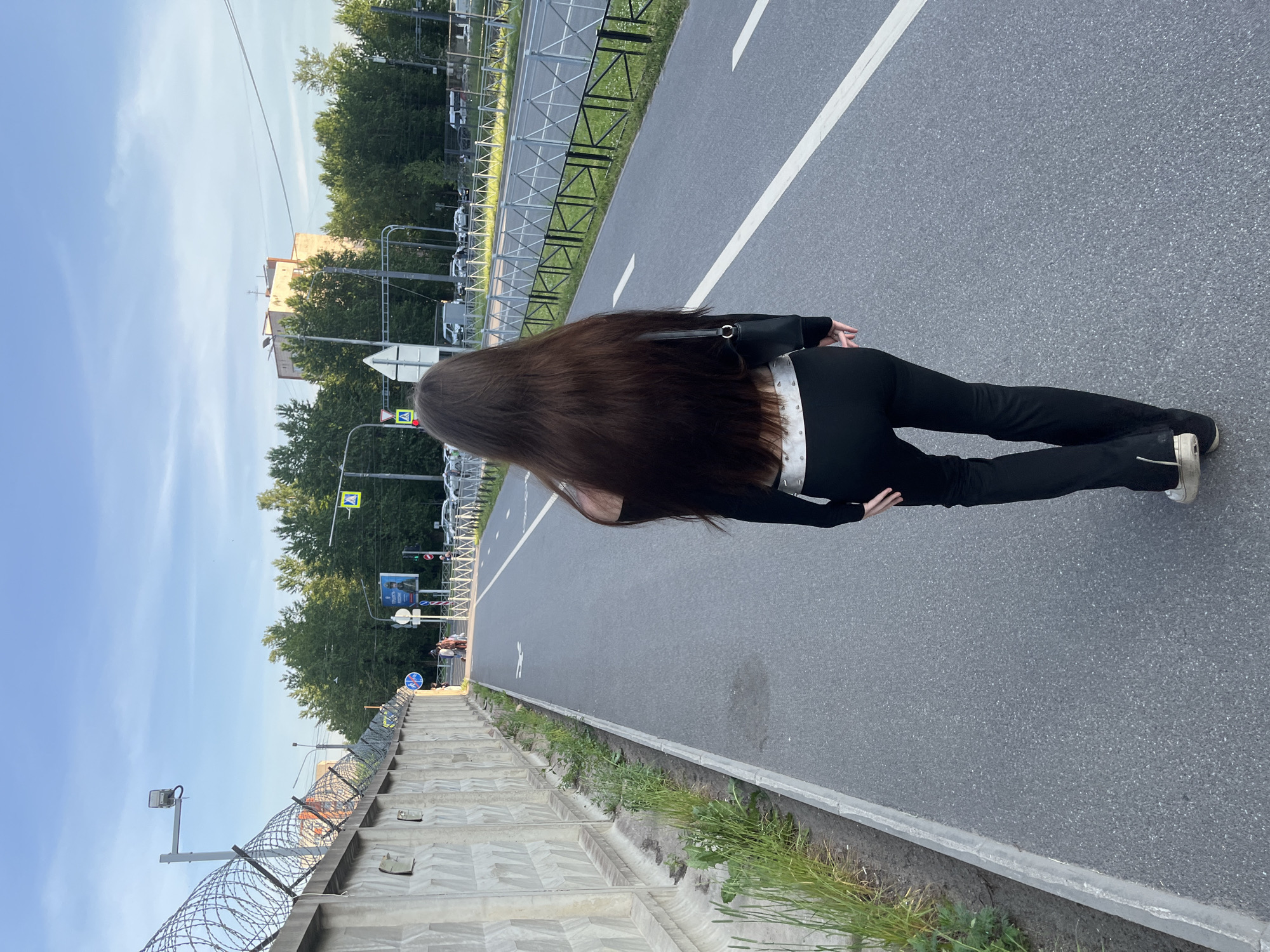 A young woman with long, dark brown hair walks away from the camera on a paved bike path. She is wearing a black off-the-shoulder top, black pants with a wide, studded belt, and light-colored sneakers. She carries a black purse on her right shoulder. The bike path is bordered on the left by a tall concrete wall topped with barbed wire, and on the right by a metal fence and a grassy area with trees beyond. In the distance, cars are visible on a road, and a multi-story building stands among the trees. The scene appears to be an urban environment in Saint Petersburg, Russia, possibly near a secured area given the fencing. The sky is partly cloudy with a blue hue.