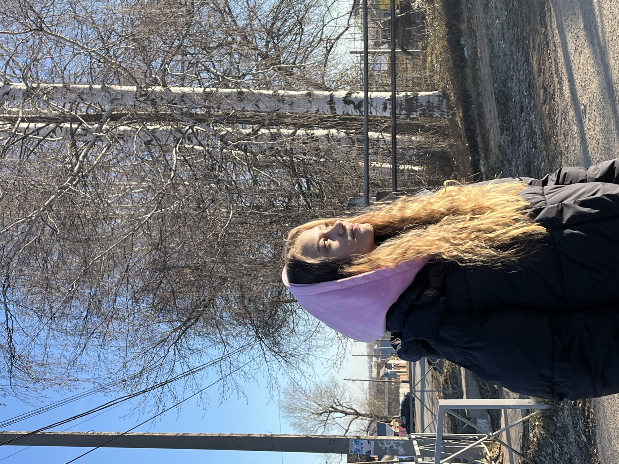 A woman with long, curly, blonde hair and a pink hood around her head is standing on a sidewalk. She is wearing a black puffy coat. To her left are several birch trees with no leaves. A gray road and sidewalk are on her right. In the background is a fence and buildings that seem to suggest the location is Фоки, Russia. Power lines cross the blue sky above. The image is rotated 90 degrees clockwise.