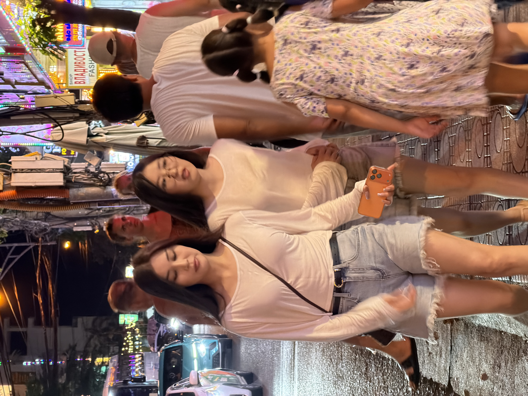 In Nha Trang Ward, Vietnam, a street scene at night captures several individuals walking along a bustling city sidewalk. In the foreground, two young women walk side-by-side. The woman on the left, with long dark hair, wears a white long-sleeved shirt and light blue denim shorts, a black belt cinching her waist. She holds an orange smartphone in her right hand. The woman next to her, also with dark hair, wears a similar white long-sleeved shirt and light brown shorts. Further behind them, a young girl with dark hair in pigtails, wearing a floral print dress, walks away from the camera. In the background, other pedestrians are visible, some appearing to be tourists. Cars, including a light blue sedan, are present on the wet street, illuminated by streetlights and neon signs that advertise shops and businesses, contributing to the vibrant nocturnal atmosphere. The street appears wet, suggesting recent rain.
