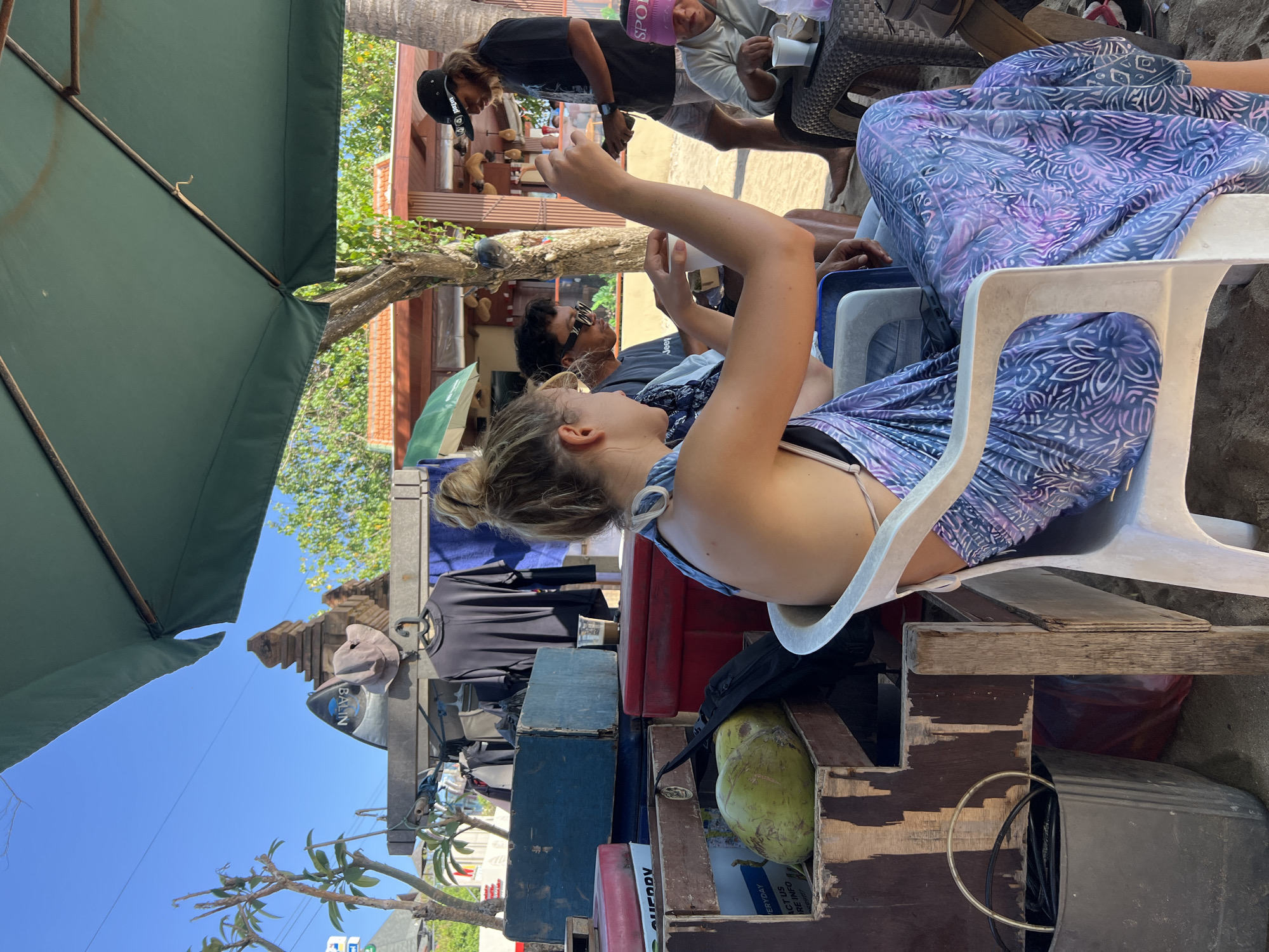 The image depicts a bustling beachside scene in Kuta, Indonesia, under a clear blue sky. The foreground is dominated by a young woman seated in a white plastic chair, her blonde hair tied up in a bun. She is wearing a purple and blue patterned sarong that drapes over her legs and a bikini top. Her arms are raised, and she holds a white cup in her right hand, appearing to be engaged in conversation or observation.
Behind her, two men are seated at a small table. One man, wearing sunglasses and a dark t-shirt, looks towards the woman. The other man, standing and wearing a black baseball cap and t-shirt, is looking down at his phone. Further to the right, another individual wearing a pink visor is seated at a table.
The setting is a beachside vendor stall, indicated by a variety of objects. A large green coconut sits in a wooden crate filled with other items, including a black backpack. Behind the seated individuals, a rack displays dark t-shirts and other clothing items. A large green umbrella provides shade over the area. In the background, traditional Balinese architecture is visible, including a temple-like structure and thatched roofs of buildings, characteristic of Kuta's landscape. The sand on the beach is visible beneath the seating area. The overall impression is a casual, lively atmosphere at a popular tourist spot.