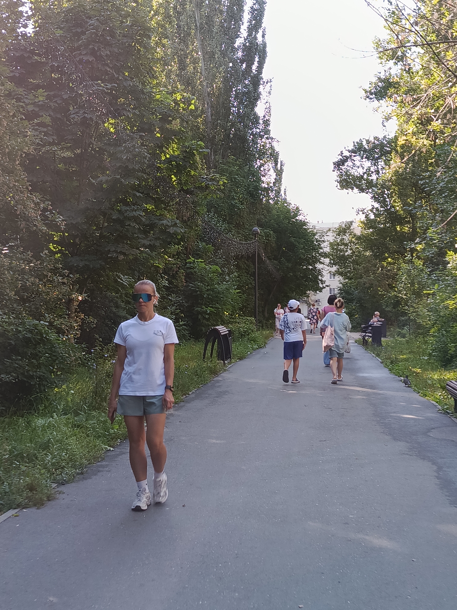 The image depicts a paved pathway in a park setting, likely in Saratov, Russia, given the architecture of a distant building. The pathway is lined with abundant green trees and foliage on both sides. Sunlight filters through the trees, creating dappled light on the path. In the foreground, a woman with blonde hair pulled back is walking towards the camera. She is wearing a white t-shirt, light-colored shorts, white athletic shoes, and large reflective sunglasses. She appears to be wearing wireless earbuds. Behind her, further down the path, several other individuals are walking away from the camera. One person in the middle distance is wearing a white t-shirt with a graphic print and blue shorts. To their right, another person is wearing a light blue t-shirt and shorts. Further back, more people are visible walking in the same direction, and a building with multiple windows can be seen in the background. Benches are present along the side of the path. The overall scene suggests a pleasant, sunny day in a public park.