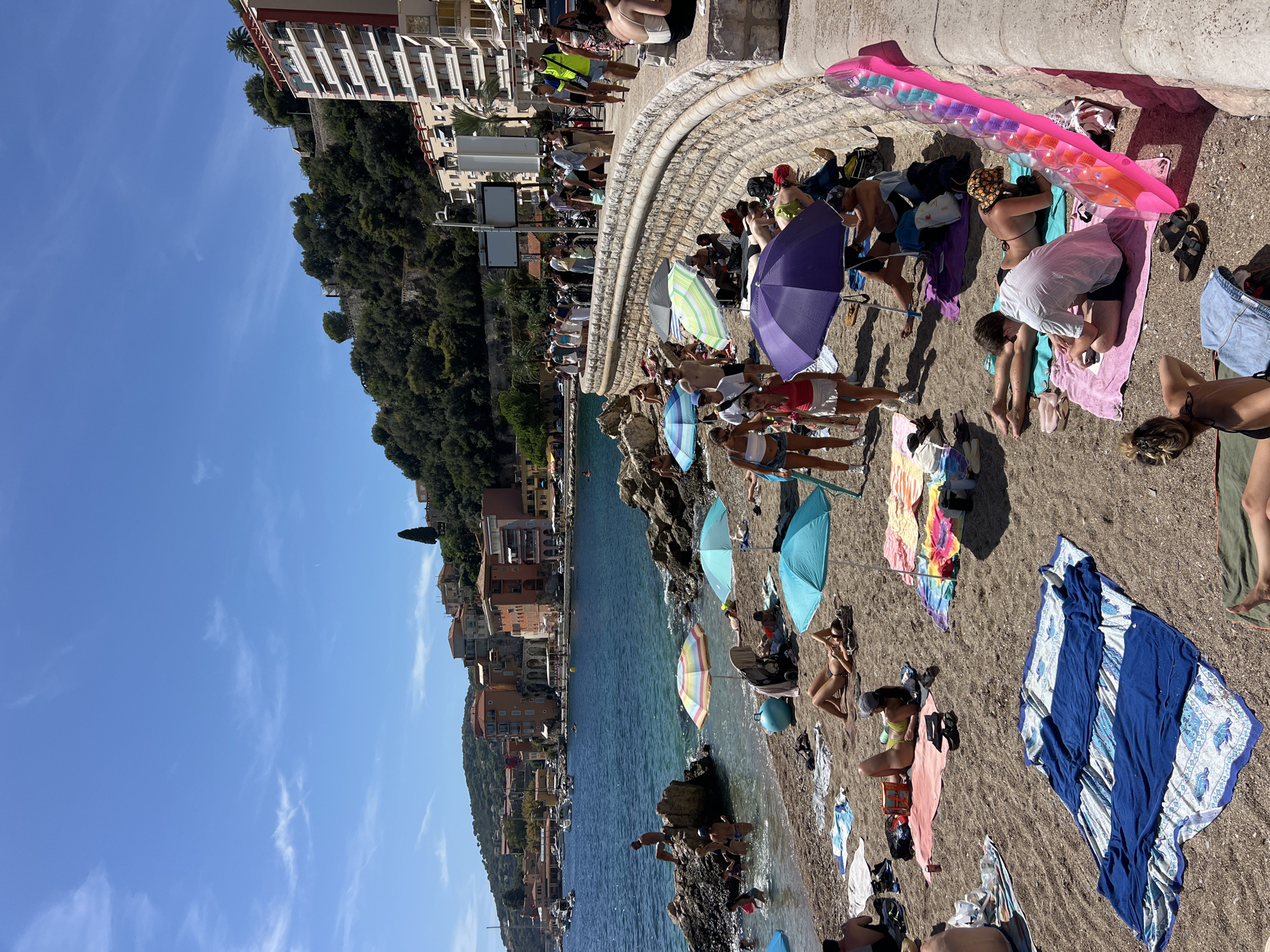 This is a daytime, eye-level shot of a crowded beach in Villefranche-sur-Mer, France. The beach is pebble and sand, with a clear blue body of water and a row of buildings on the hillside behind it. The people are sunbathing, swimming, or walking along the beach. Many are under beach umbrellas. The water is calm, and there are boats in the distance. There are various colorful beach towels and umbrellas spread out on the sand. A stone wall runs along the edge of the beach promenade, with people standing or walking on it. The weather is sunny and clear, with a few wispy clouds in the sky.