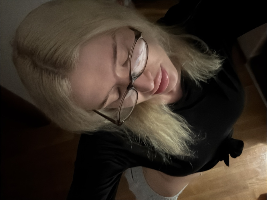 A blonde-haired individual, presumed female, is depicted from a high-angle perspective, looking downward. The subject wears dark-framed eyeglasses with reflections on the lenses, a black long-sleeved top, and light grey lower garments. A distinct light source, originating from below the subject, illuminates her face and reflects on her lower lip and the eyeglass lenses. The immediate environment includes light-colored wooden floorboards visible on the lower left. The background is dark and indistinct. The specific location within Budva, Montenegro, cannot be determined from the visual information provided.