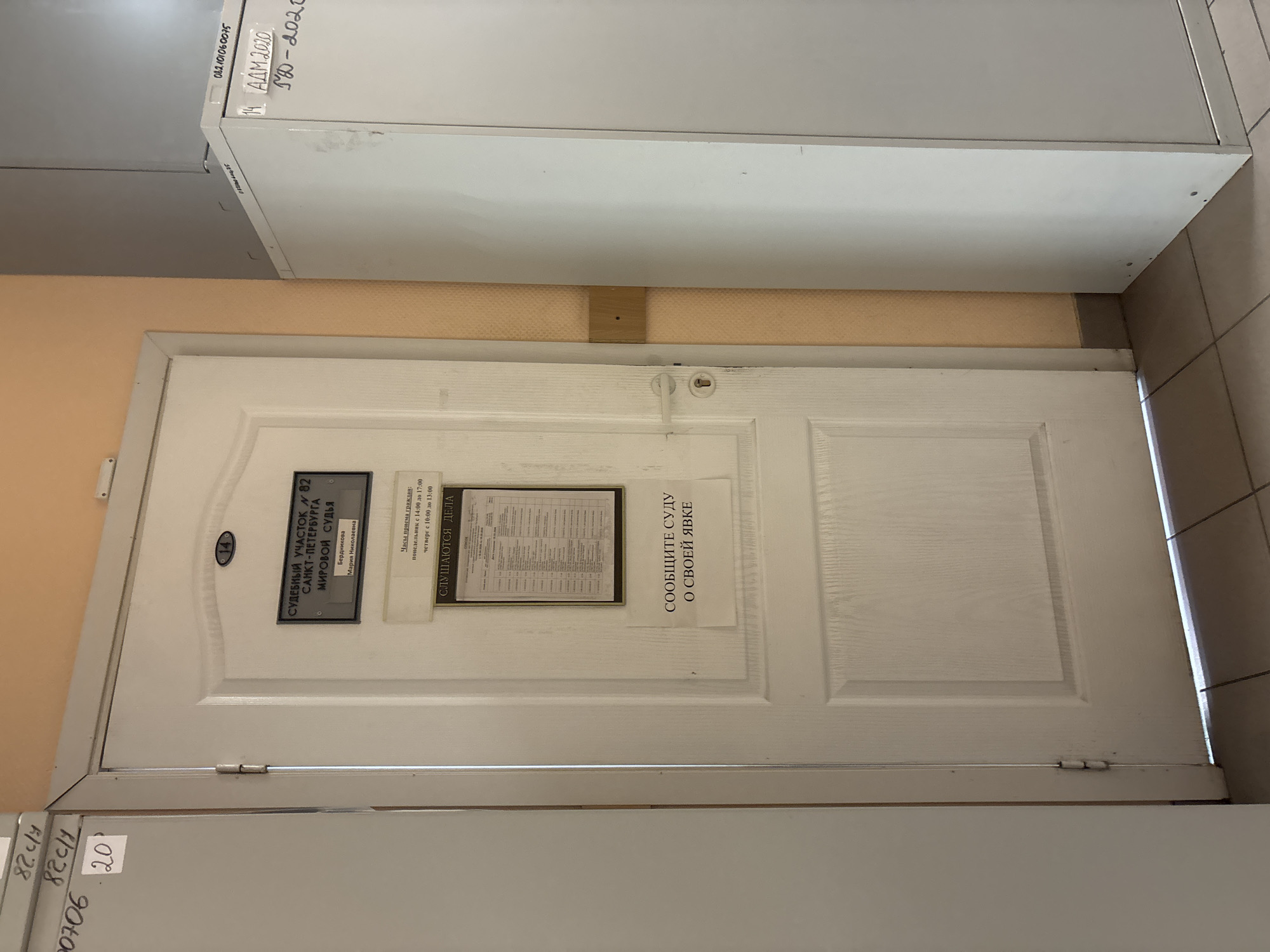 The image shows a white door with three notices on it. The notice on the left identifies the location as Judicial District No. 82 of Saint Petersburg, Russia, presided over by Magistrate Maria Nikolaevna Berdnikova. The middle notice lists the hours of citizen reception as Mondays from 2:00 PM to 5:00 PM and Thursdays from 10:00 AM to 1:00 PM, and also states “Cases are being heard.” The notice on the right states “Inform the court of your appearance.” Above the door are white shelves. The lighting is natural. The door appears to be in an institutional setting, possibly a courthouse.