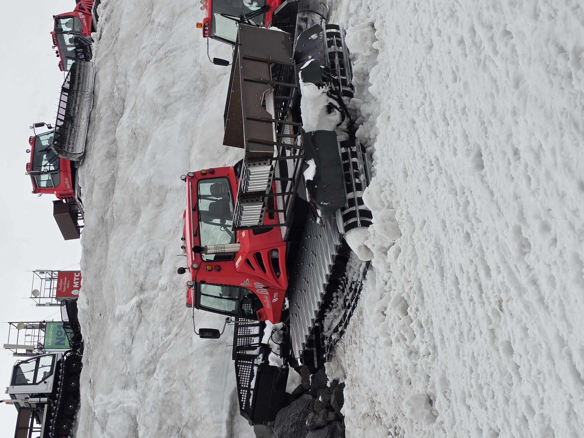 A bright red, tracked snow groomer, identified as a PistenBully 600, is parked on a snow-covered slope. The machine has a large plow in front and a bed for carrying equipment or passengers in the rear. Other snow groomers and ski lifts are visible in the background on a snowy mountainside, suggesting a ski resort setting. The sky is overcast. In the background, a sign identifies the location as "Elbrus", the highest peak in Russia, and mentions "Terskol", a village at the foot of Mount Elbrus. There are no people visible in the image.