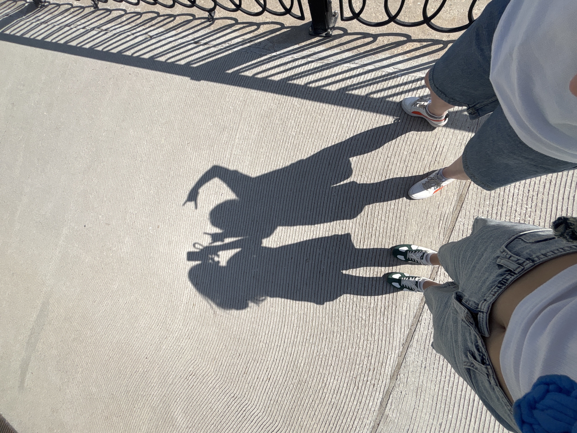 The image shows an overhead shot of two people standing on a concrete surface, likely a sidewalk or walkway. Sunlight casts shadows of the individuals, revealing one person holding a camera and gesturing with a raised finger. The people are wearing casual clothing including shorts and light-colored shirts. Their footwear includes white sneakers with colored accents and green and white sneakers. A metal railing casts linear shadows on the concrete. The overall setting suggests an outdoor urban environment. There is no indication of the location.