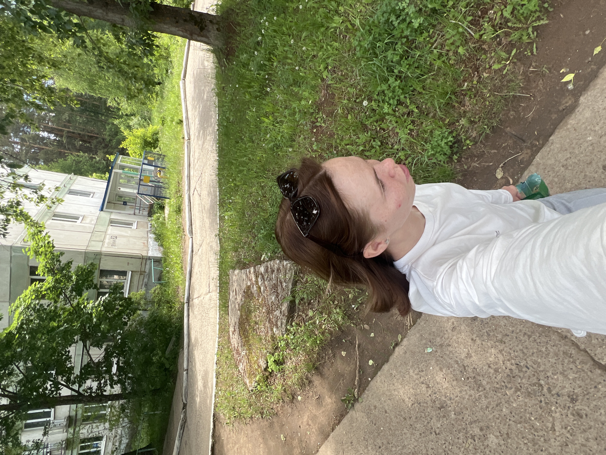 A young woman is standing outdoors in what appears to be a residential area. She is wearing a white long-sleeved shirt and has sunglasses on her head. In the background, there is a concrete path next to a grassy area and a building with trees in front of it. The location is Chaikovsky, Russia, as indicated in the prompt.