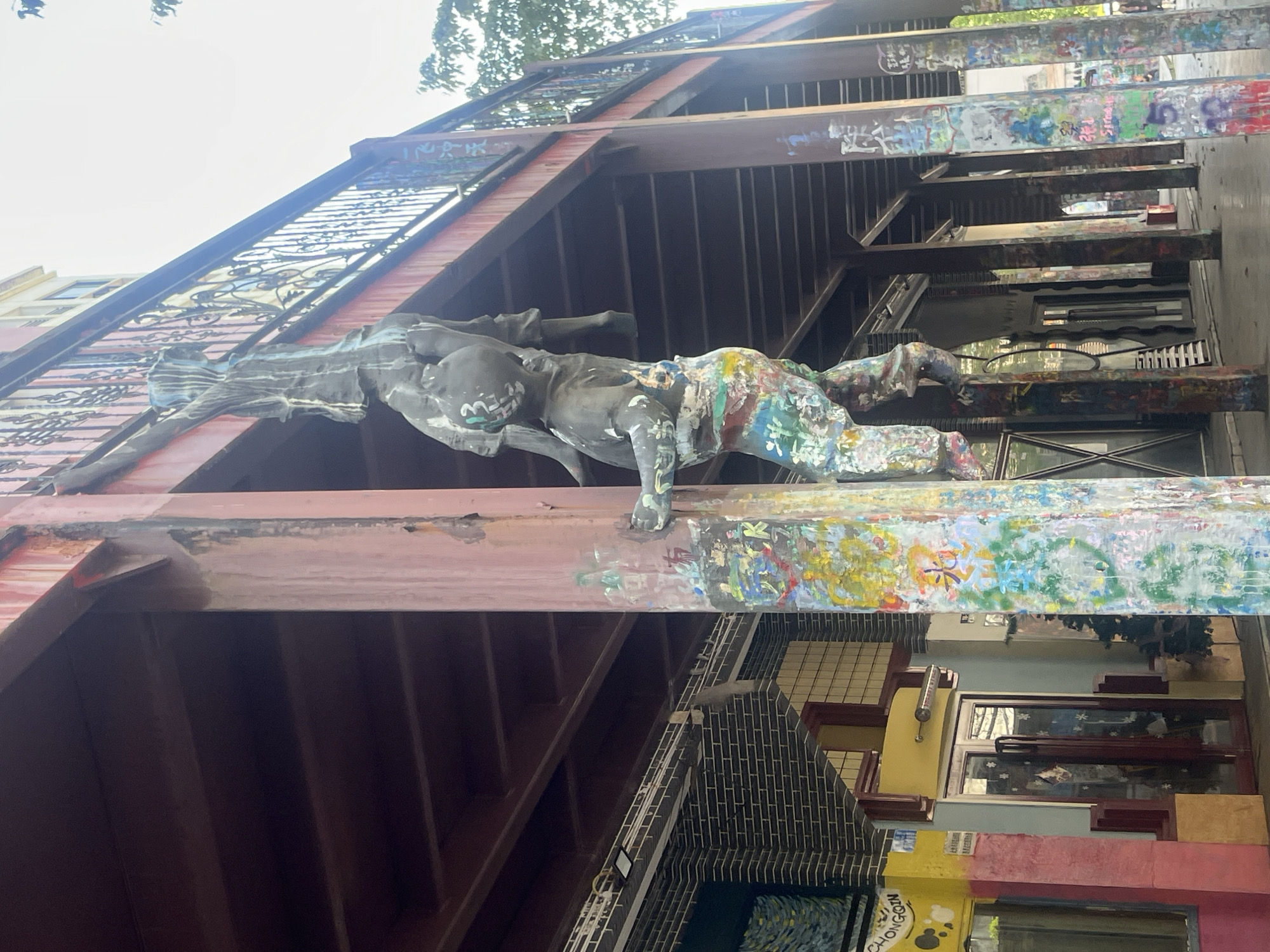 The image depicts a building in Jiulongpo District, China, likely an art installation or a building facade with artistic elements. The most striking feature is a sculpture of a reclining figure mounted on a steel beam structure. The figure is painted with a mix of colors on the lower body, contrasting with a dark or unpainted upper body. The building's architecture is multi-layered, including exposed steel beams, staircases, windows, and walls decorated with various artistic designs and graffiti. The overall aesthetic suggests a creative or bohemian environment. The image is taken from a low angle, looking up at the building and sculpture.