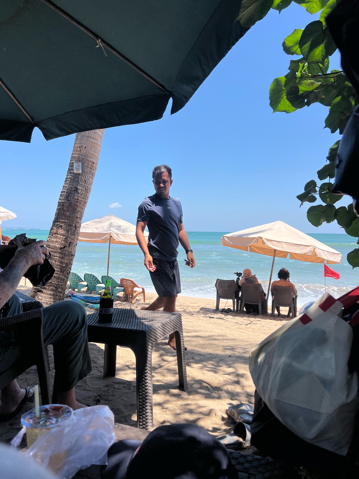 On a bright, sunny day in Kuta, Indonesia, a man stands on a sandy beach, looking towards the left of the frame. He is wearing a dark blue t-shirt and dark shorts. Behind him, the turquoise ocean meets a clear blue sky, with gentle waves rolling onto the shore. Several beach umbrellas and chairs are visible, suggesting a relaxed atmosphere. To the left, a palm tree stands tall, and a person is seated in a chair, holding a black object. A table with a bottle of beer and a drink with a straw is in the foreground. In the distance, under a beige umbrella, two people are seated, facing the ocean. A red flag with a white symbol is planted in the sand to the right. The overall scene depicts a typical day at the beach in Kuta, characterized by sunshine, the ocean, and people enjoying leisure time.