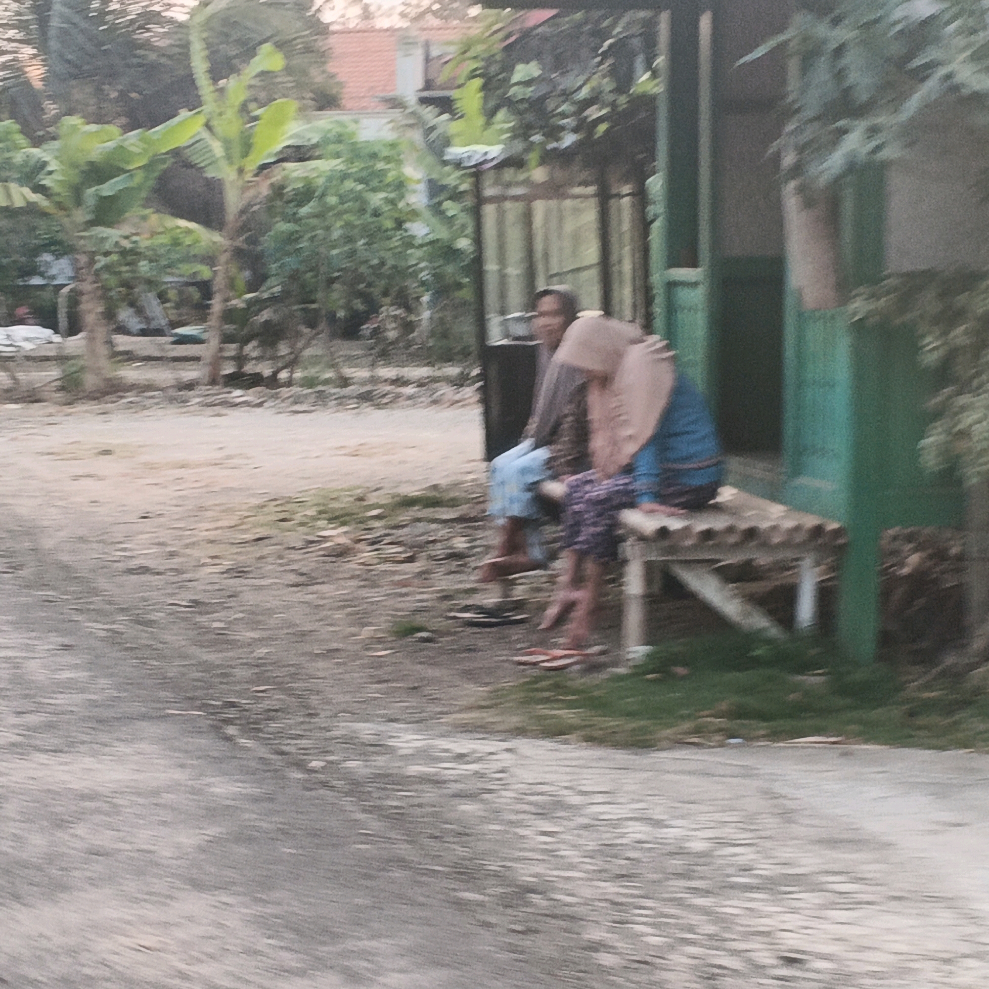 In Gentasari, Indonesia, two individuals are seated on a wooden bench situated outdoors. The bench is constructed of corrugated wood and supported by legs. To the right of the bench, a green structure with vertical panels is visible. The ground in front of the bench is dirt, and a paved road runs across the foreground. Lush vegetation, including banana trees and other greenery, surrounds the area, with a building roof visible in the background. The scene appears to be in a rural or semi-rural setting.