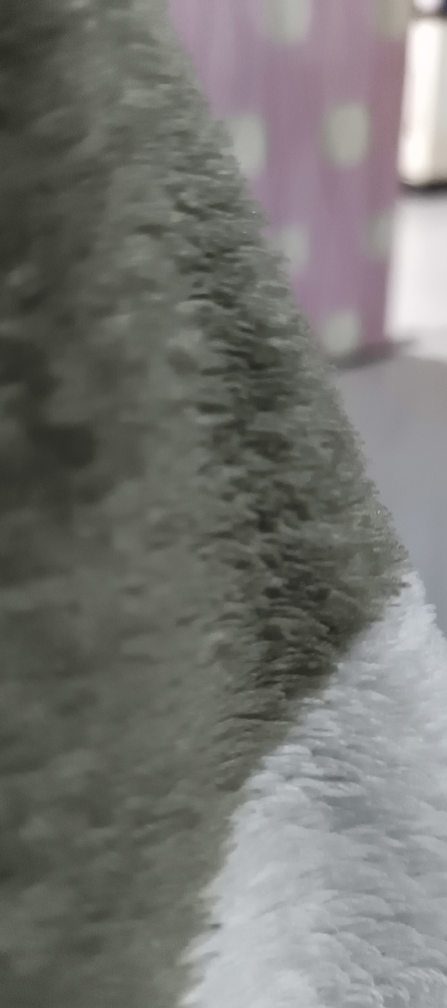 The image is a close-up, abstract shot of a textured surface, possibly fabric or fur, with a soft, plush appearance. The dominant colors are shades of gray and white, with some blurred pink and white circular patterns visible in the background. The texture appears fluffy and uneven, with individual strands or fibers distinguishable. The lighting is soft, creating subtle shadows and highlights that emphasize the texture. The background is out of focus, suggesting a shallow depth of field. There is no discernible indication of the city of Muaro Jambi, Indonesia, or any specific location within it. The image is devoid of any people, objects, or actions that would provide context for its origin or setting.