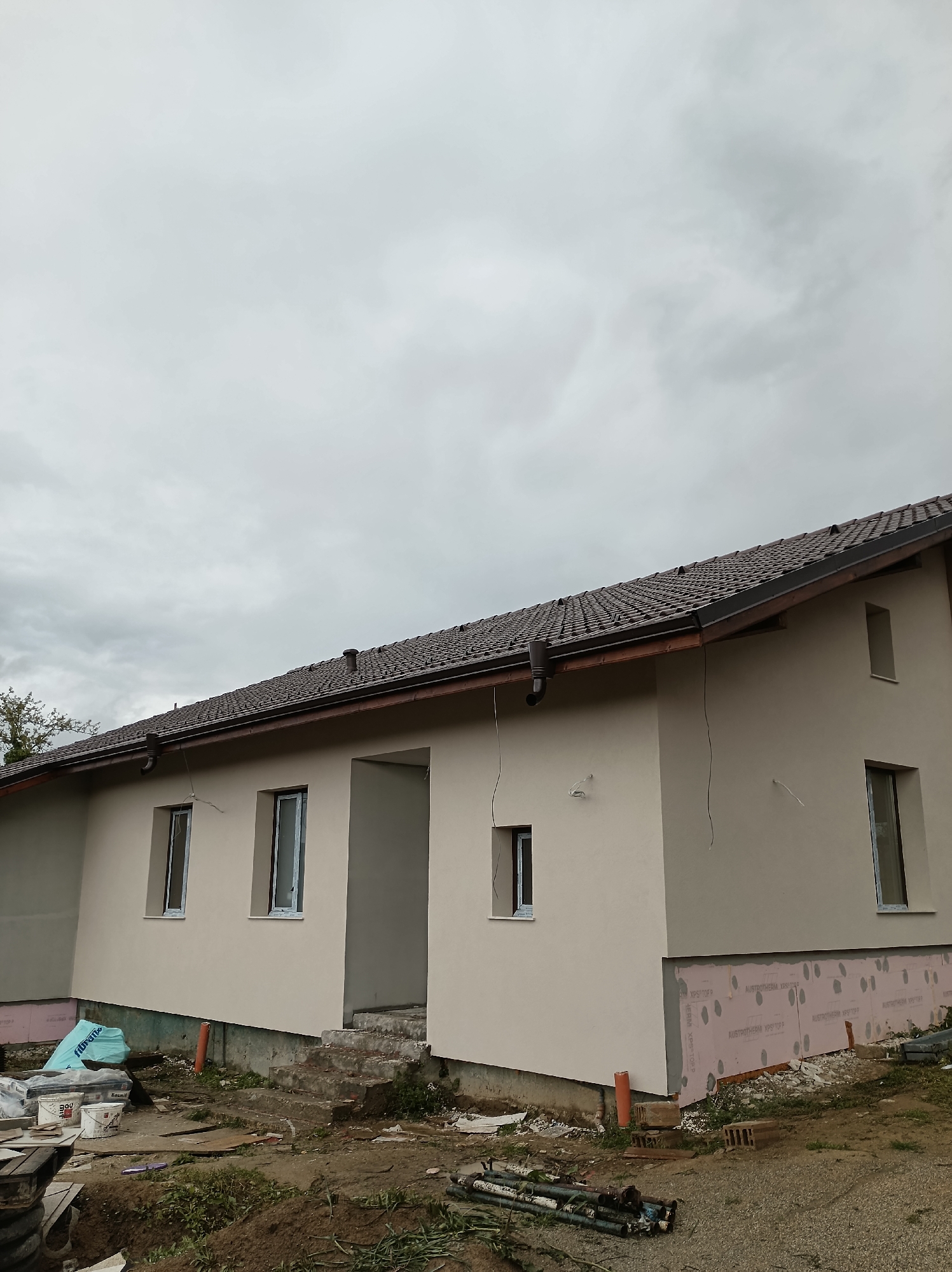 The image shows a single-story house in Mihaylovtsi, Bulgaria, under a cloudy sky. The house has a brown tiled roof and a light beige exterior with several windows. Some windows are open, while others appear to be still under construction. The lower portion of the house's exterior is covered with pink insulation panels. The ground around the house is dirt and gravel, with construction debris, including pipes, buckets, and building materials, scattered around. The steps leading to an open doorway appear to be unfinished concrete. A few trees are visible in the background.