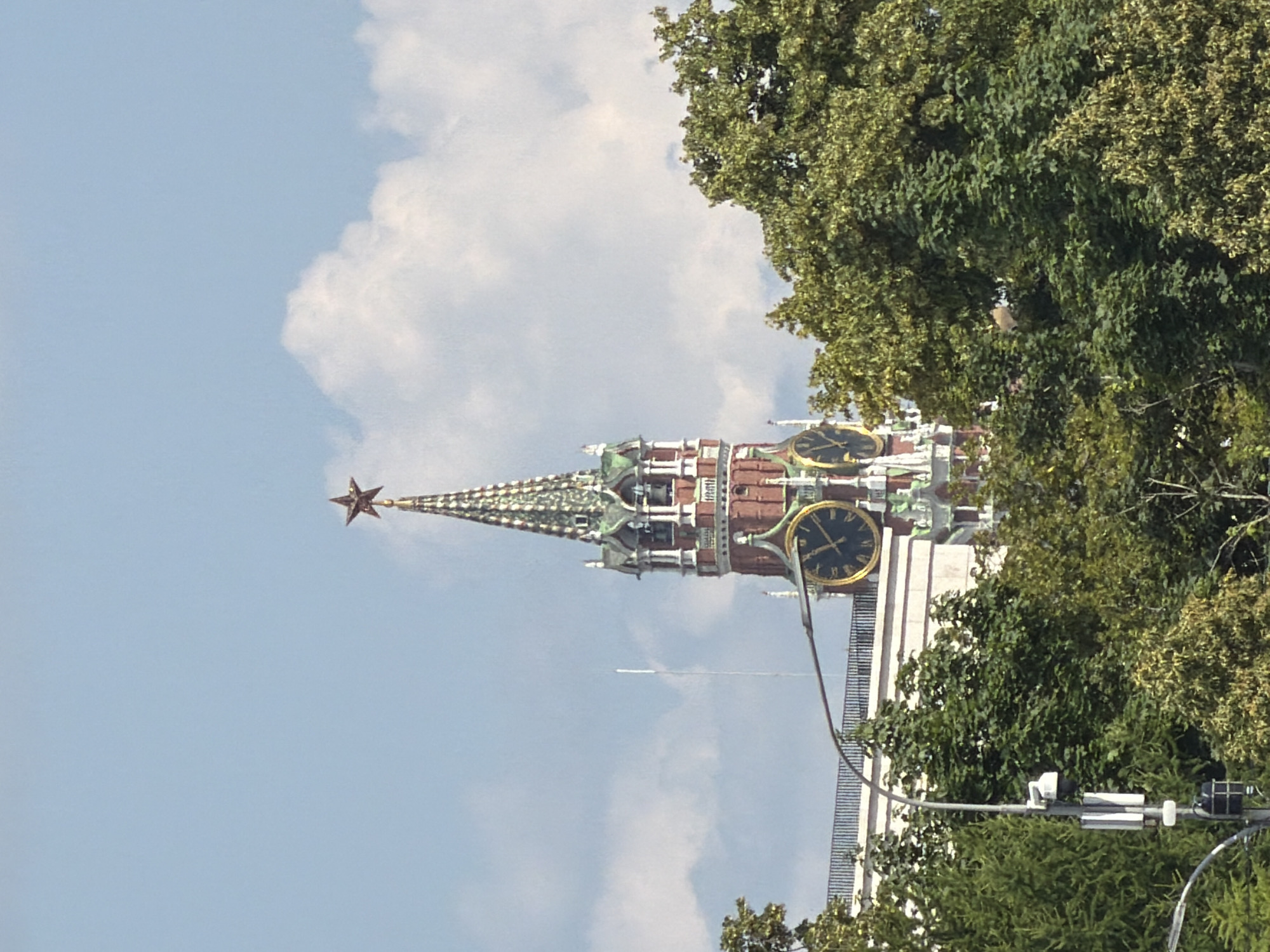 The image captures a view of the Spasskaya Tower, a prominent clock tower on the eastern wall of the Moscow Kremlin, Russia. The tower, constructed of red brick with green accents and a golden spire topped by a red star, dominates the upper portion of the frame. Two large, circular clocks are visible on the tower. The tower is partially obscured by lush green trees in the foreground and mid-ground. A white railing and the top of a building are visible behind the trees and below the tower. A lamppost with a security camera is also present in the lower left of the image. The sky above is a clear blue with scattered white clouds. There are no people or discernible activity in the image.