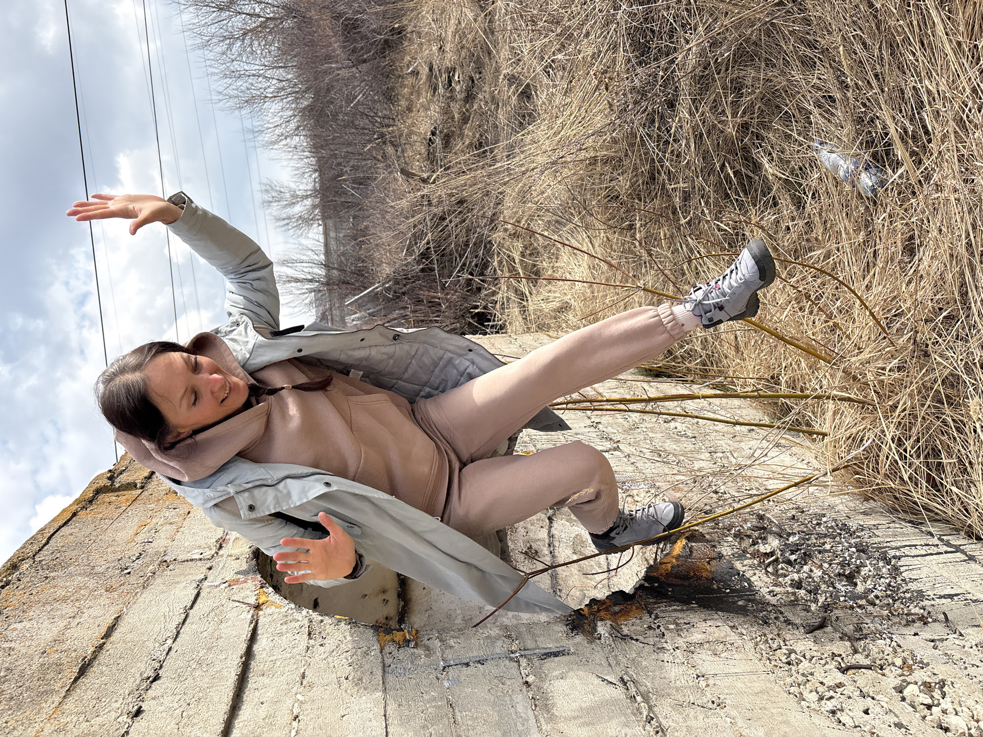 A woman is posing for a photograph in Ulyanovsk, Russia. She is reclining on a concrete structure, extending her arms outward. She is wearing a light gray jacket, a light brown hoodie, light brown sweatpants, and gray sneakers. Behind her is a patch of dry, brown vegetation. Above her, telephone wires are visible against a cloudy sky. A plastic bottle is visible amidst the vegetation. The concrete structure appears damaged, with exposed rebar and crumbling edges.