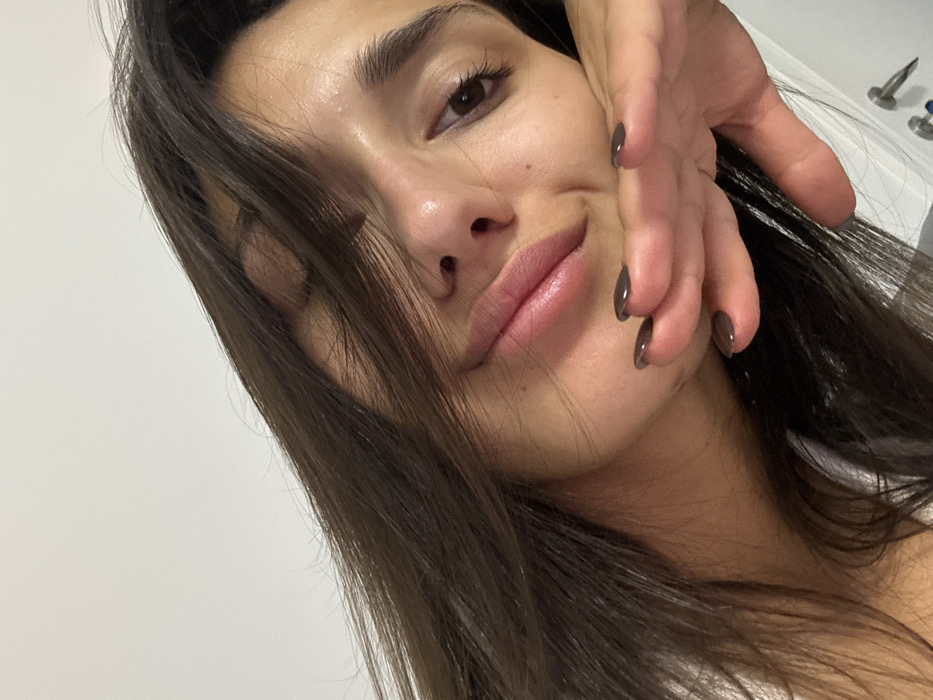 A woman with dark hair, prominent eyebrows, and fair skin poses close to the camera. One of her hands is near her face, fingers extended with dark nail polish. Her expression is a slight smile, and she's looking at the camera. The background is a plain white wall, with some indistinct objects barely visible. The lighting accentuates her facial features.