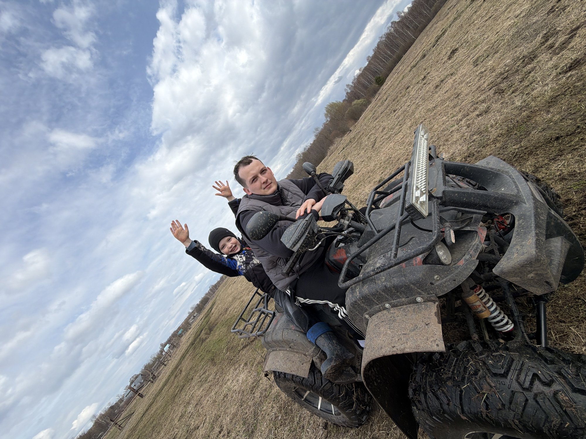 A father and child are on a four-wheeled all-terrain vehicle (ATV) in a field near Кемуль, Russia, on a cloudy day. The adult male, wearing a dark jacket and vest, sits on the ATV, operating it, and looking at the camera. A younger child, wearing a dark jacket and a beanie, sits behind him, waving. The ATV is heavily soiled with mud, indicating recent off-road use. The field consists of brown grasses and is partially visible, with trees on the horizon. The sky is overcast, with a mix of clouds covering most of the frame. The ATV is angled towards the camera, showcasing its front end and tires.