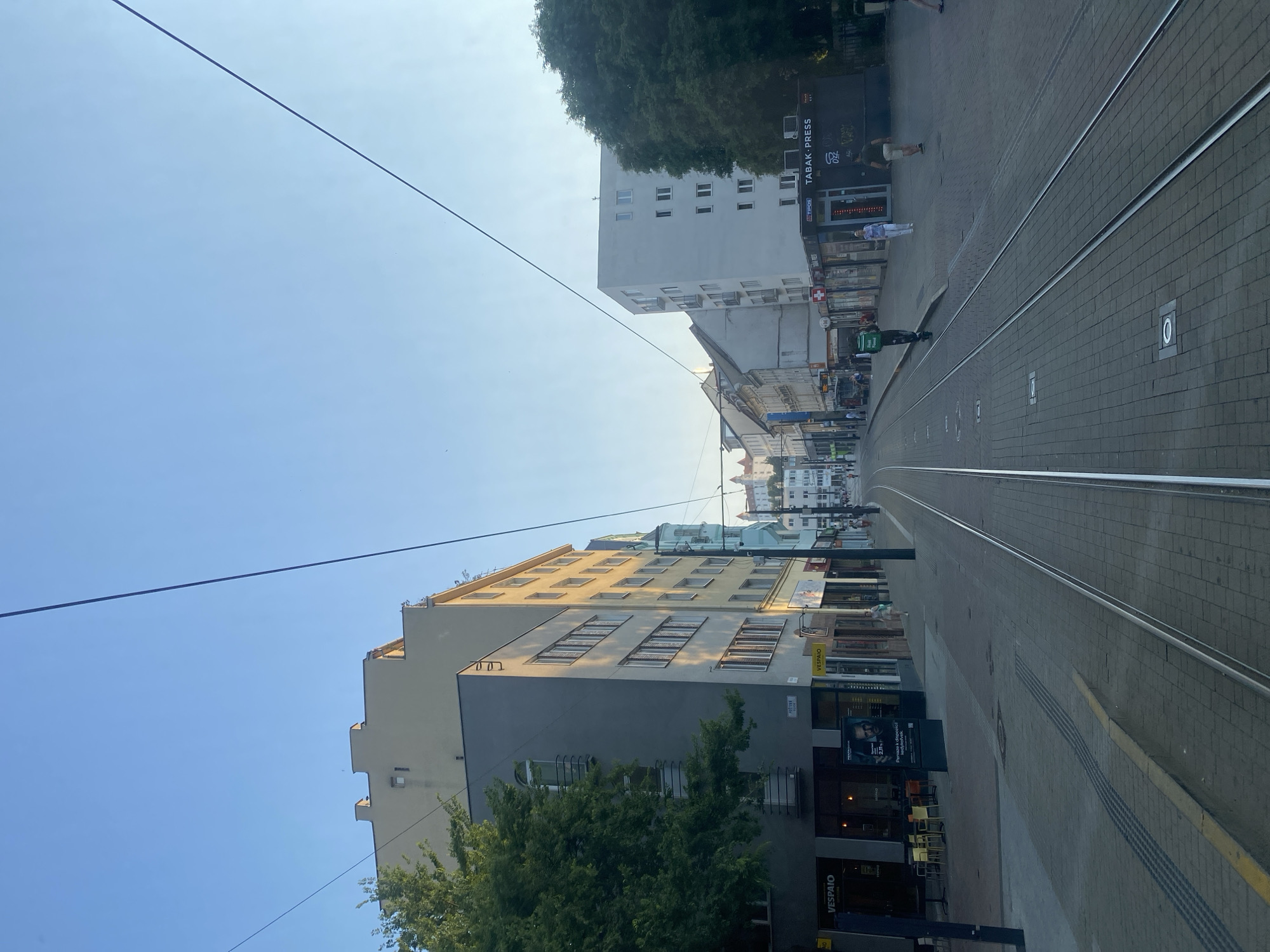 The image shows a street scene in Bratislava, Slovakia. The perspective is from a moving tram or similar vehicle, looking forward along the tram tracks which dominate the foreground. The tracks curve to the right, leading the eye down a street lined with multi-story buildings.
On the left side of the street, several buildings are visible. The closest one is a modern building with a light grey facade and dark grey lower section, with trees in front. Further down the street, a taller building with a pale yellow facade and numerous windows is prominent. To its left and further back, a white building with a simple, modern design is visible, partially obscured by trees.
On the right side of the street, the buildings are less distinct but appear to be older structures with shopfronts at ground level. One shop sign reads "TABAK-PRESS". Another sign appears to be "VESPAIO". There are also indications of outdoor seating for cafes or restaurants.
Several people are visible on the sidewalks, appearing as small figures in the distance. One person is wearing a green bag, possibly a delivery person. The sky is clear and blue, suggesting a sunny day. The lighting indicates it is likely daytime, perhaps late afternoon given the angle of the shadows. The overall impression is of a typical urban environment with active street life.