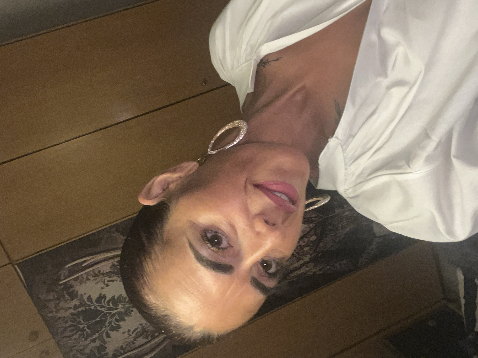 A woman with dark hair, wearing large hoop earrings and a white blouse, poses for a selfie. She is indoors, with wooden cabinets visible in the background. The image does not provide any information about the location in the city of Konz, Germany.