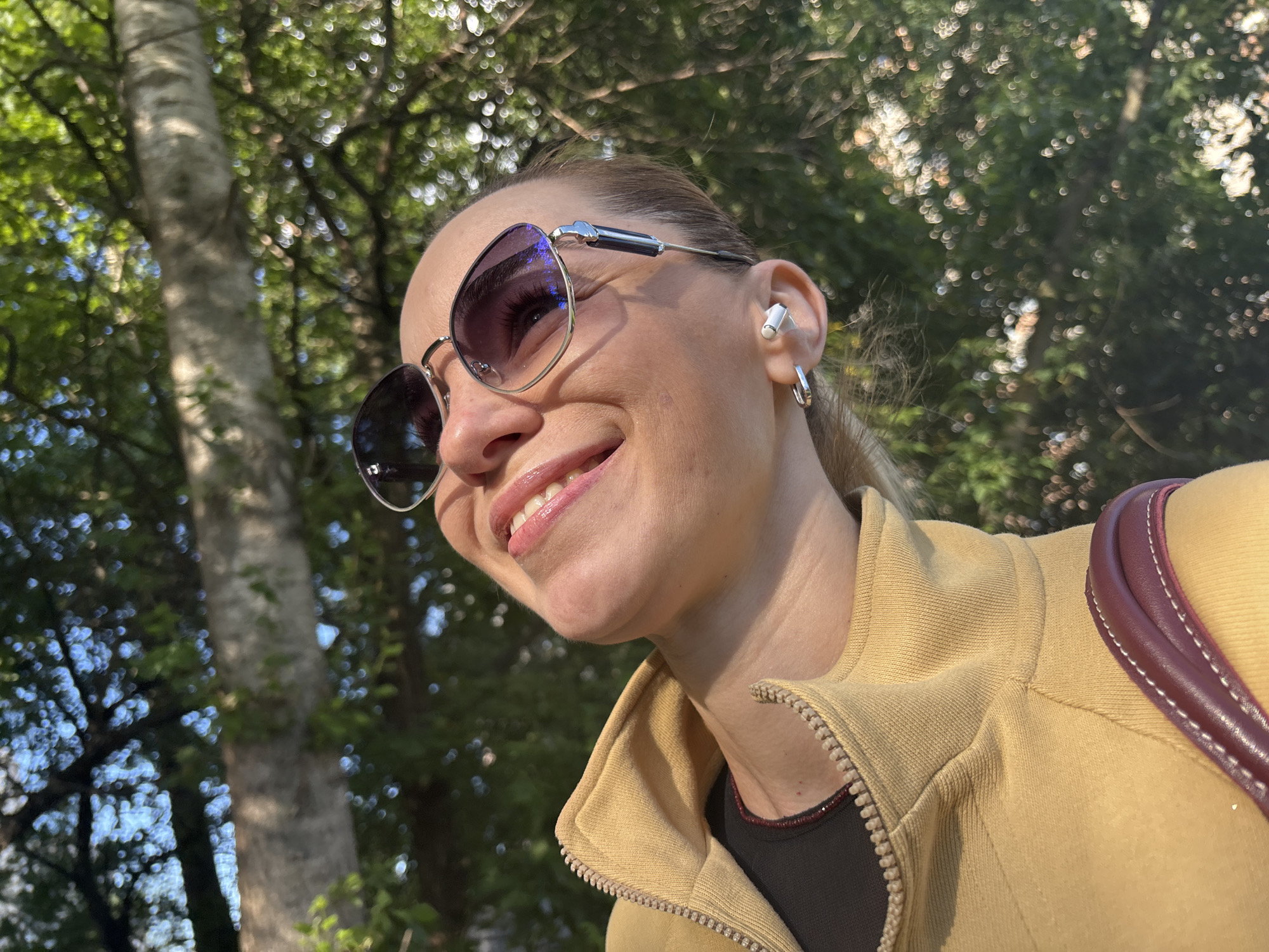 A close-up, eye-level outdoor shot captures a woman's face from the chin up. She is wearing large, reflective sunglasses, and a white wireless earbud is visible in her right ear, along with a silver hoop earring. Her mouth is open in a smile, revealing her teeth and glistening lips. She has on a yellow zippered jacket over a dark garment, and a maroon strap of a bag rests on her right shoulder. The background is blurred, showing green foliage and tree trunks, suggesting a park or natural setting within an unknown city in Russia. The lighting indicates it is daytime, with sunlight filtering through the leaves.