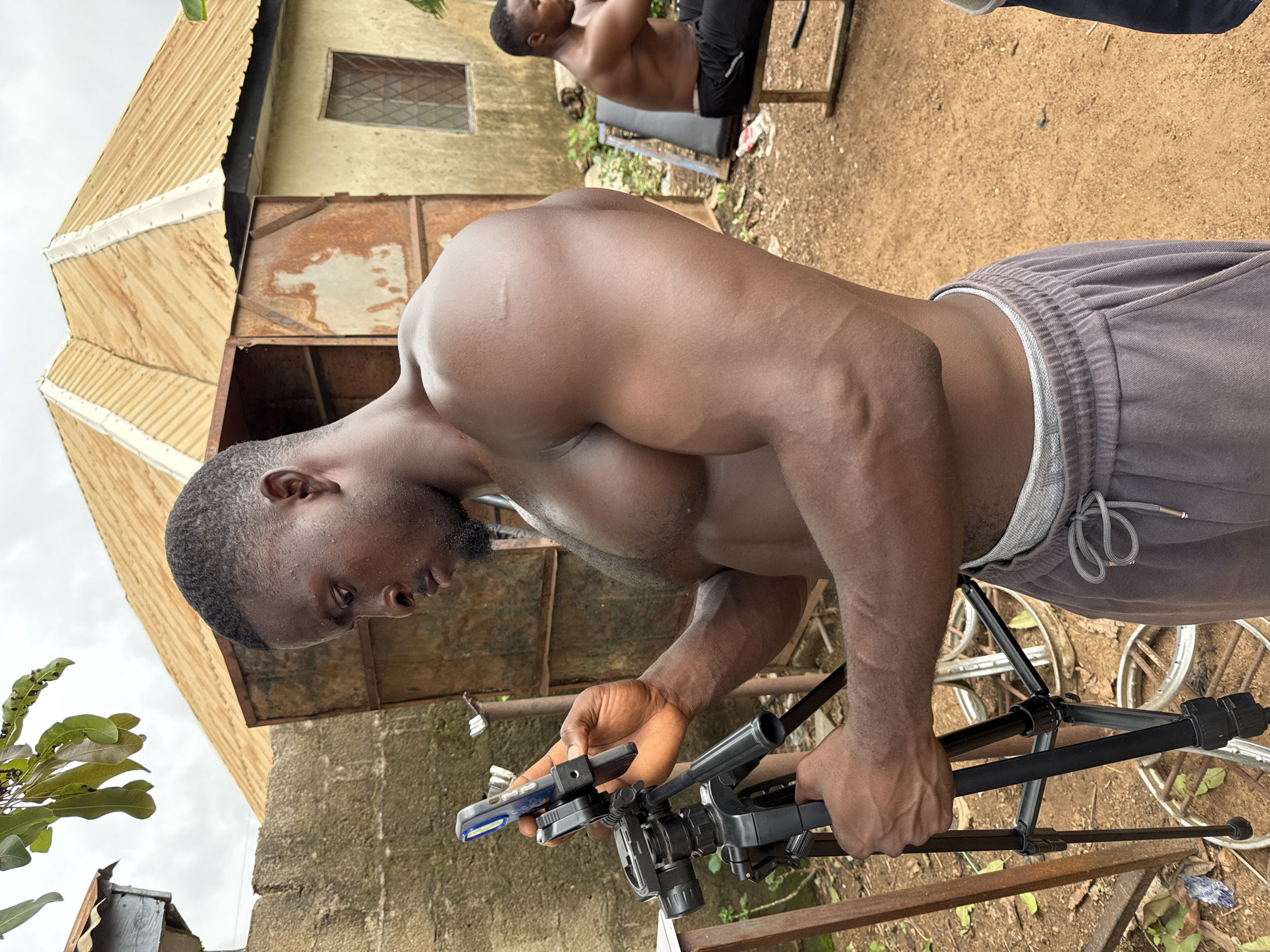 The image depicts a muscular, shirtless man with dark skin holding a smartphone mounted on a black tripod. He is wearing grey sweatpants. The man is focused on the phone. In the background, another man is visible exercising on a bench. The environment appears to be an outdoor space in Akwanga, Nigeria. The background includes a building with a corrugated metal roof, rust-colored panels, and a small window. The ground is dirt. There are bicycle wheels scattered on the ground. The leaves are on the bottom-left, in the corner of the image.